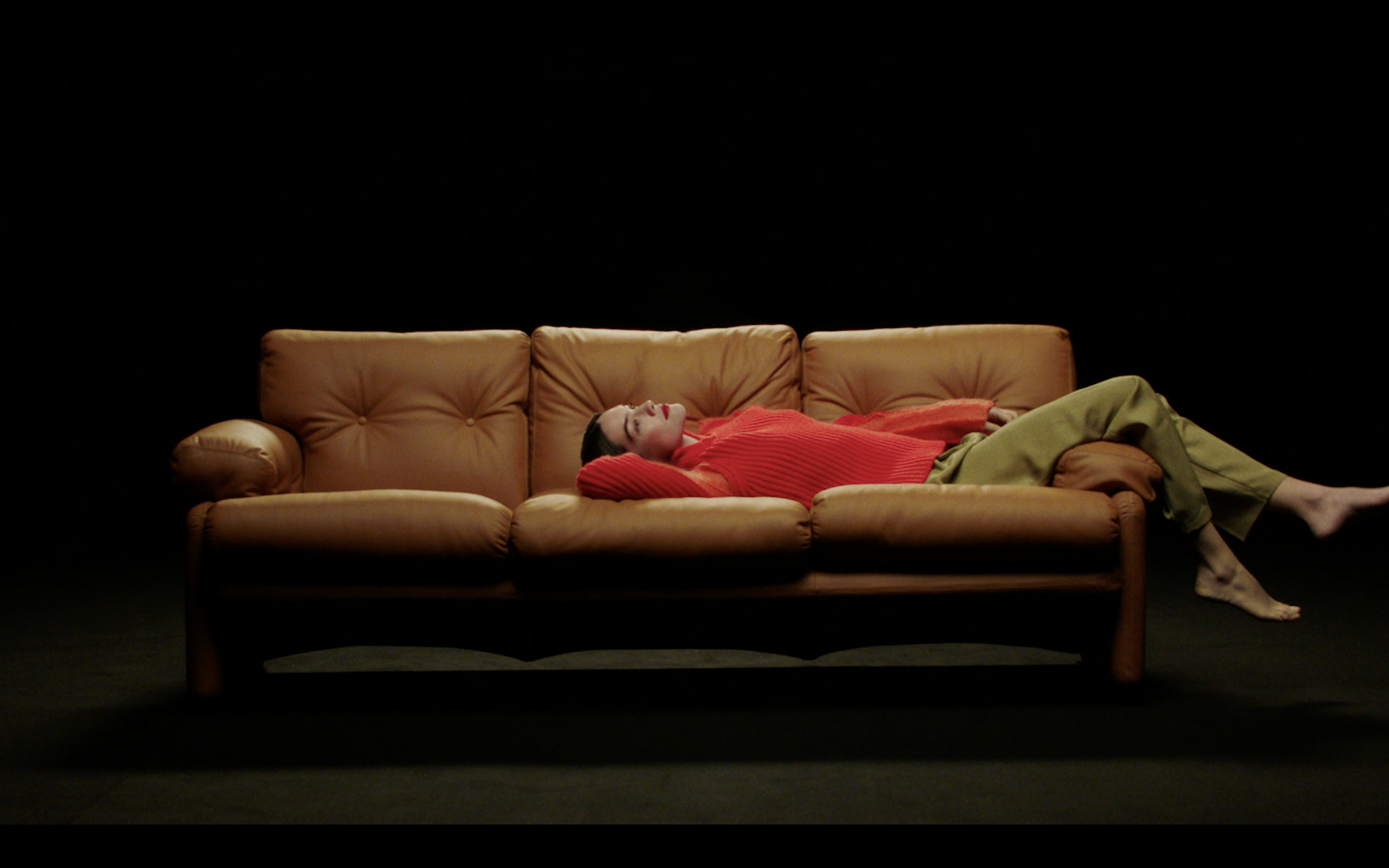 Donna sdraiata su un divano marrone, con sfondo nero, che indossa un maglione rosso e pantaloni verdi.