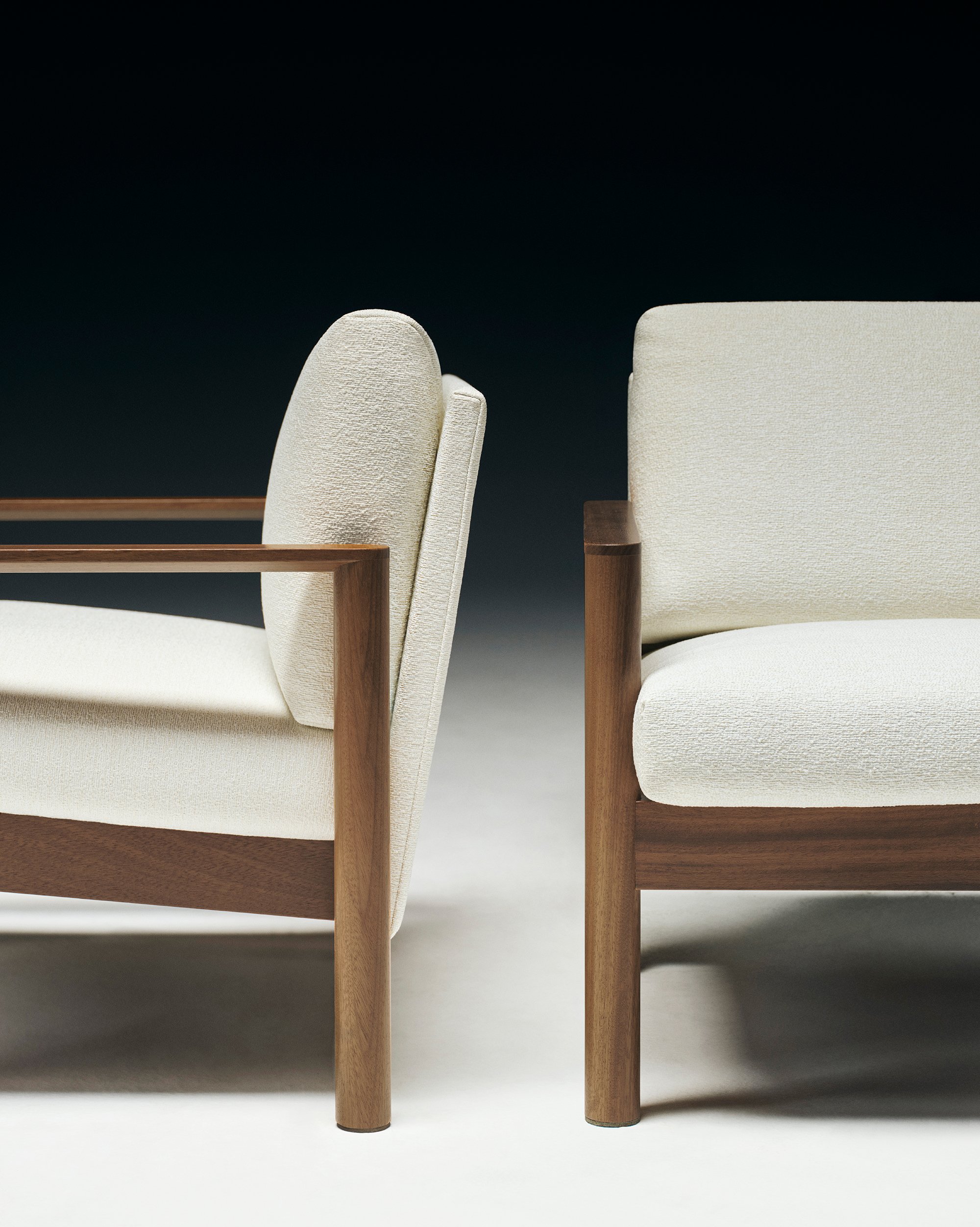 Due sedie con struttura in legno e rivestimento beige, una con braccioli e l'altra senza, disposte su uno sfondo scuro.