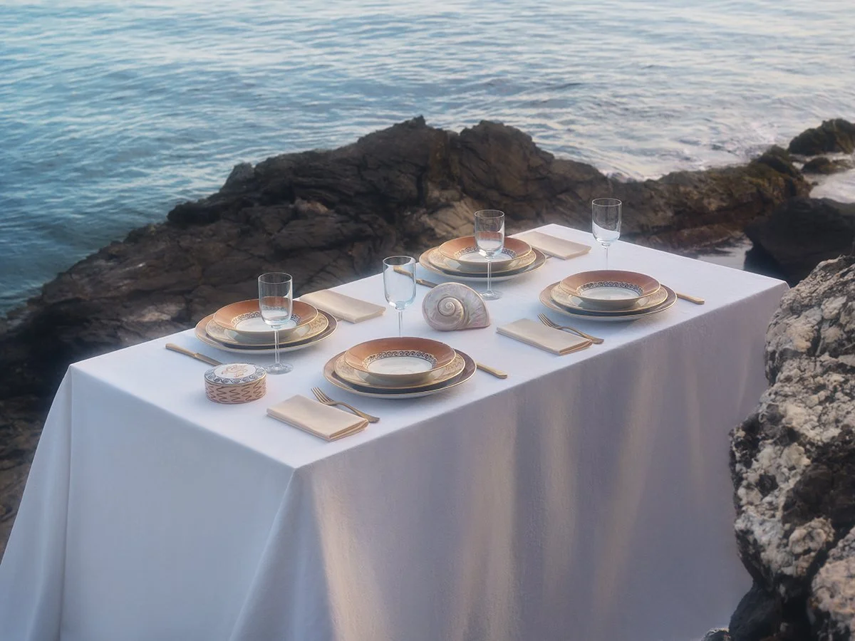 Tavola apparecchiata con piatti, bicchieri e posate, situata vicino alla riva del mare con rocce.