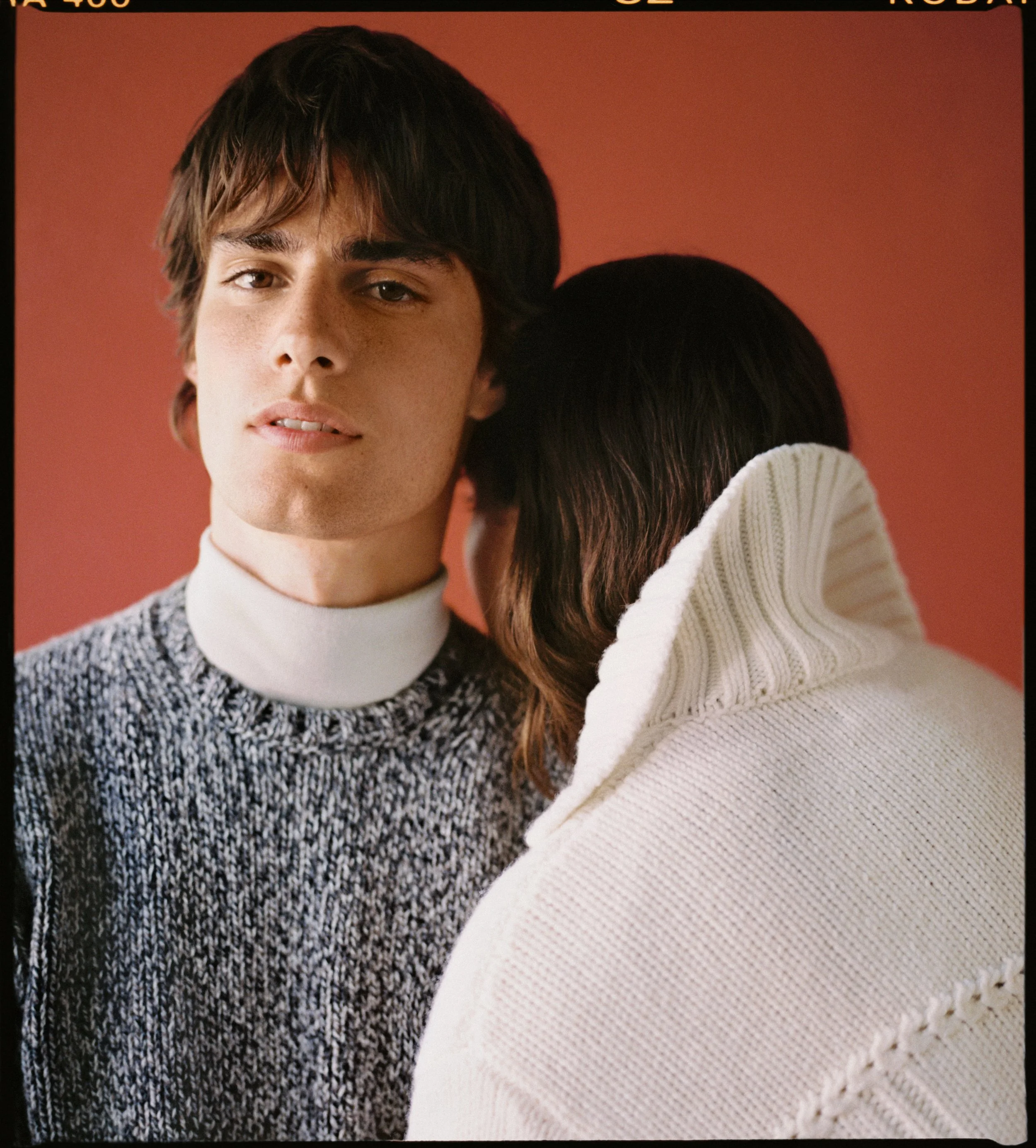 Un giovane uomo con capelli castani e occhi chiari sorride leggermente, indossa un maglione a maglia e una camicia bianca sotto, mentre una donna con capelli castani scuri coperti da un cappotto bianco in lana si appoggia alle sue spalle con il viso rivolto verso di lui contro uno sfondo arancione.