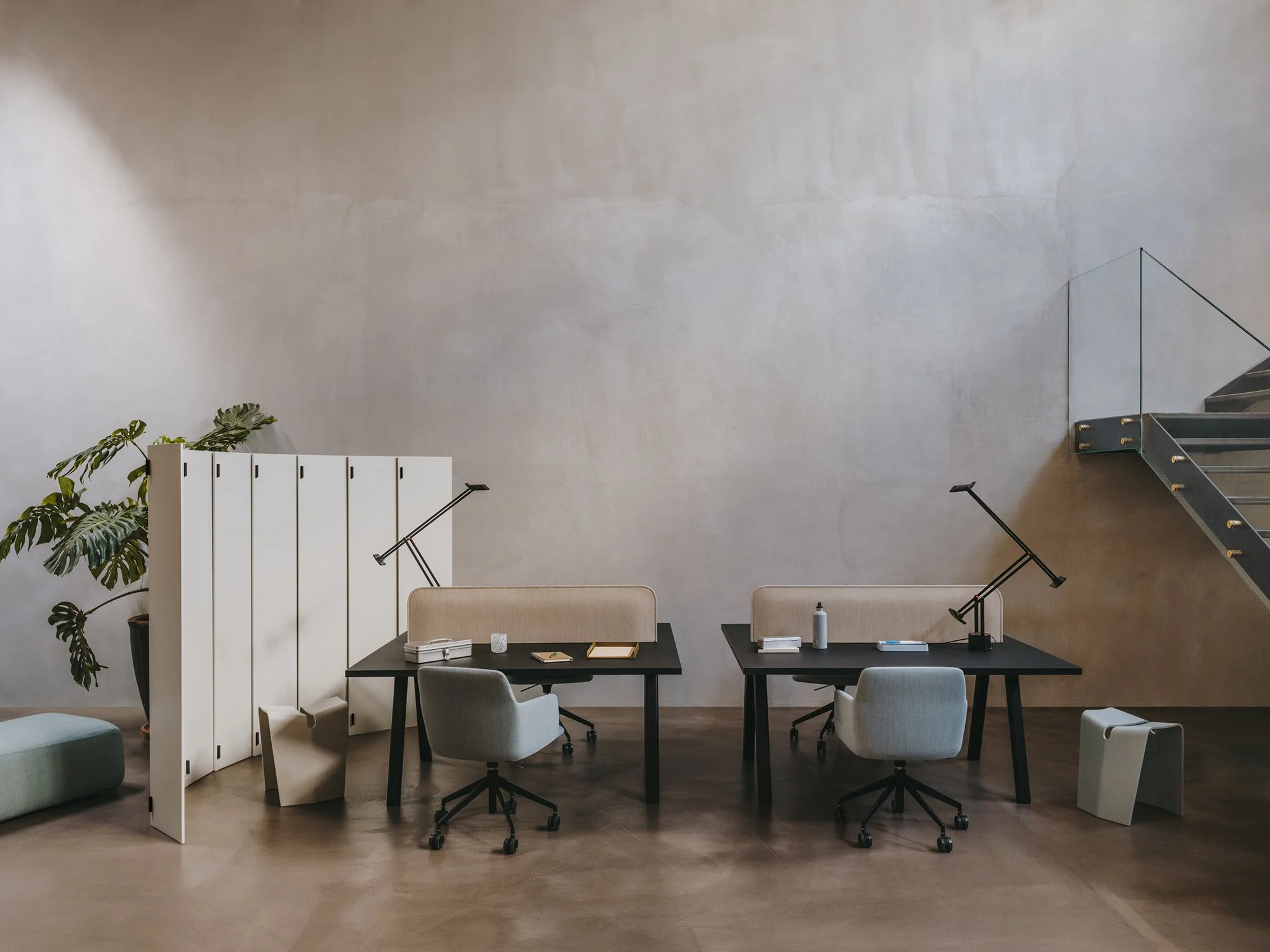 Uno spazio di coworking con due scrivanie nere, sedie grigie, lampade da tavolo nere, una parete divisoria bianca, una pianta e una scala metallica con vetro.