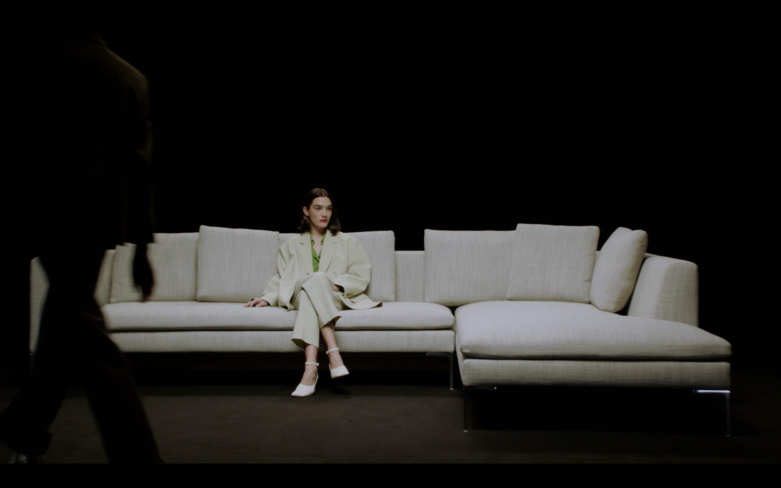 Una donna seduta su un divano chiaro, indossa un completo beige con scarpe bianche, in un ambiente con sfondo nero e un'altra persona sfocata attraversa l'immagine a sinistra.