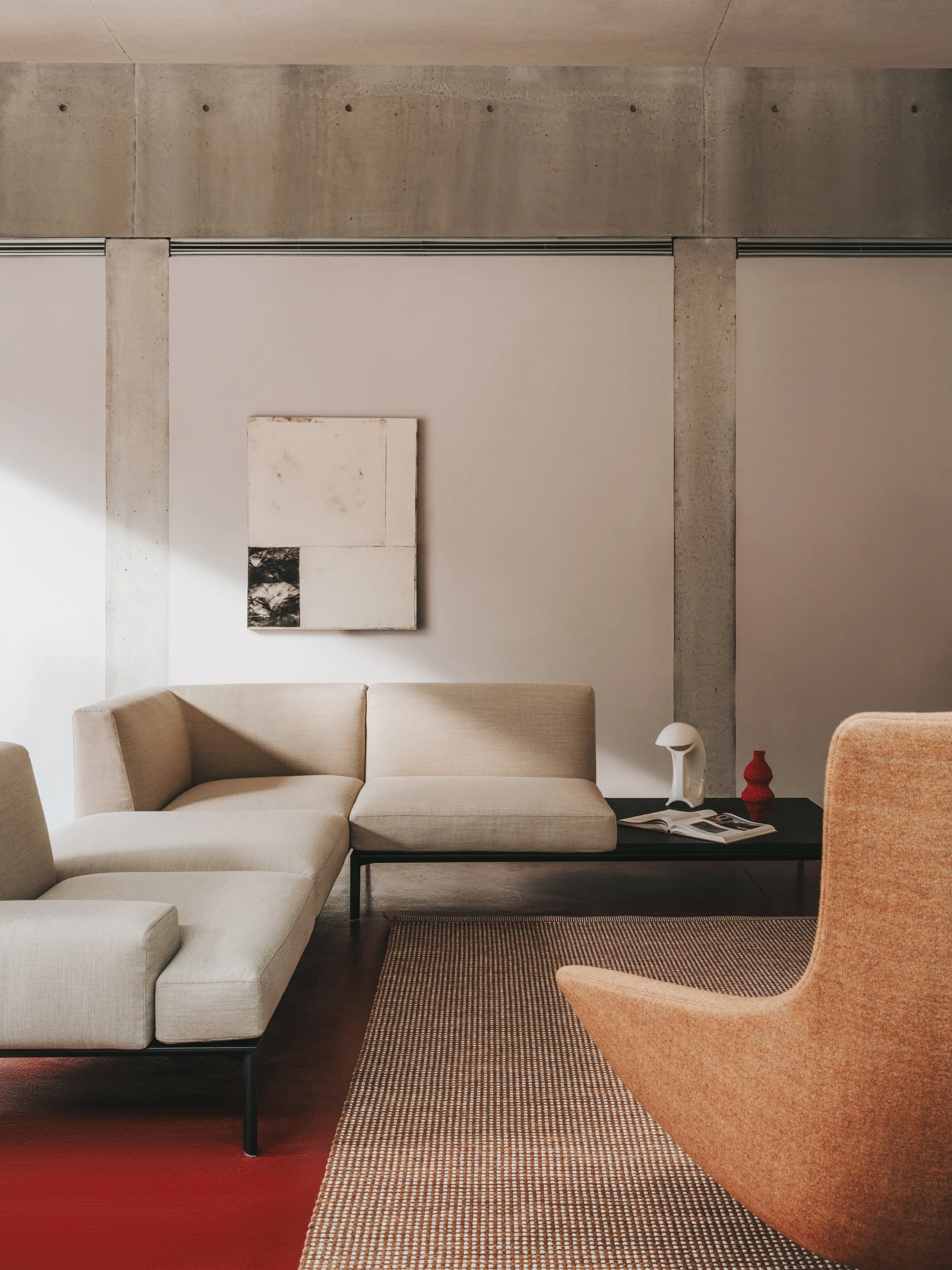 Interno di un soggiorno con divani beige, una sedia arancione, un tavolino nero con riviste e decorazioni, pareti in cemento con un quadro astratto.