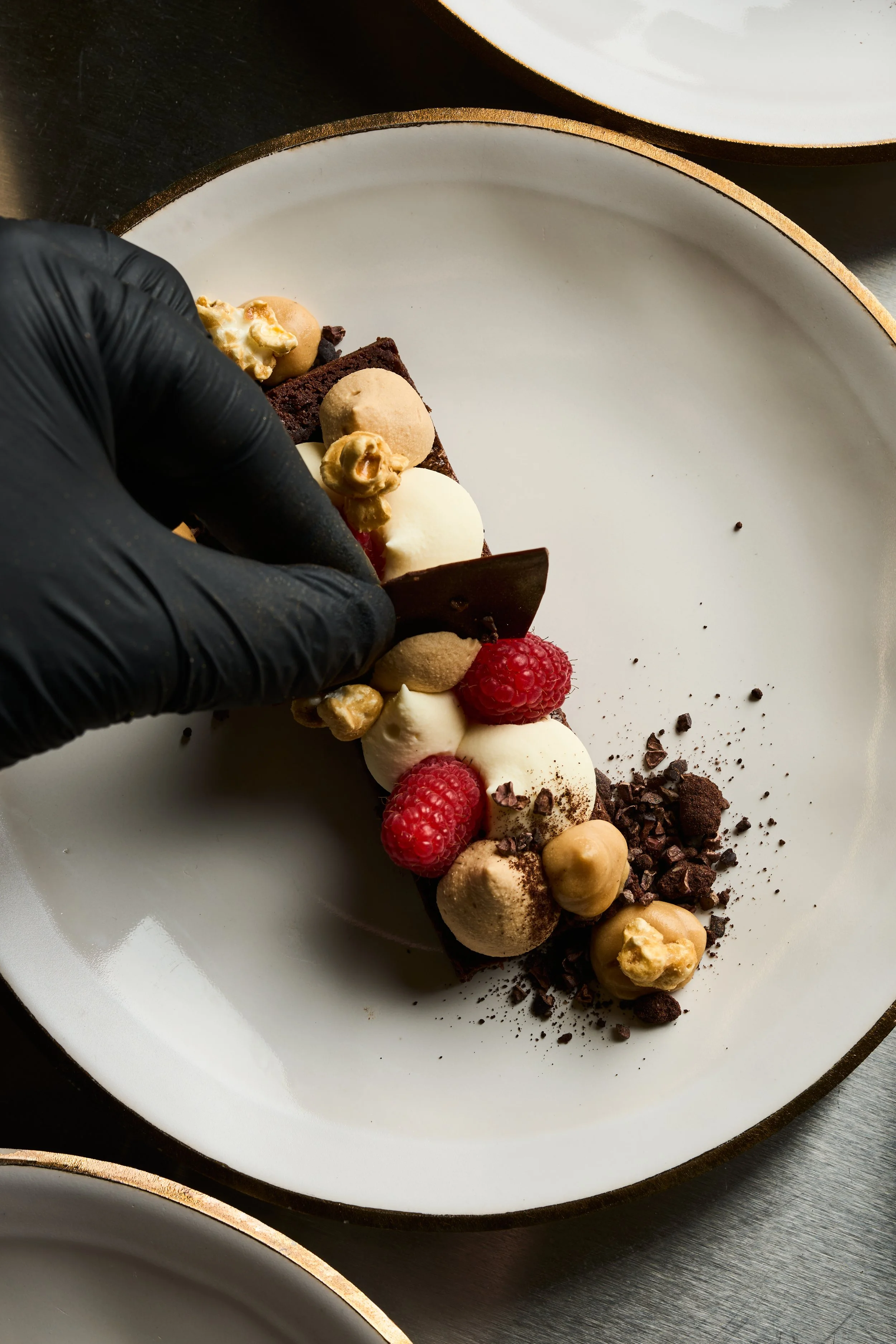 Ein Küchenchef oder Konditor dekoriert ein mehrschichtiges Schokoladendessert mit Beeren, Waffeln und Schokoladenstückchen auf einem weißen Teller.