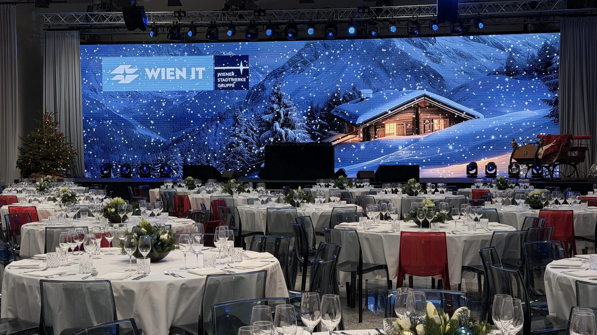 Festlich gedeckte Tische in einem Saal mit großer Bühne und winterlichem Bergmotiv im Hintergrund, Weihnachtsbaum auf der linken Seite, Konfetti und Lichter auf den Tischen, festliche Dekorationen.