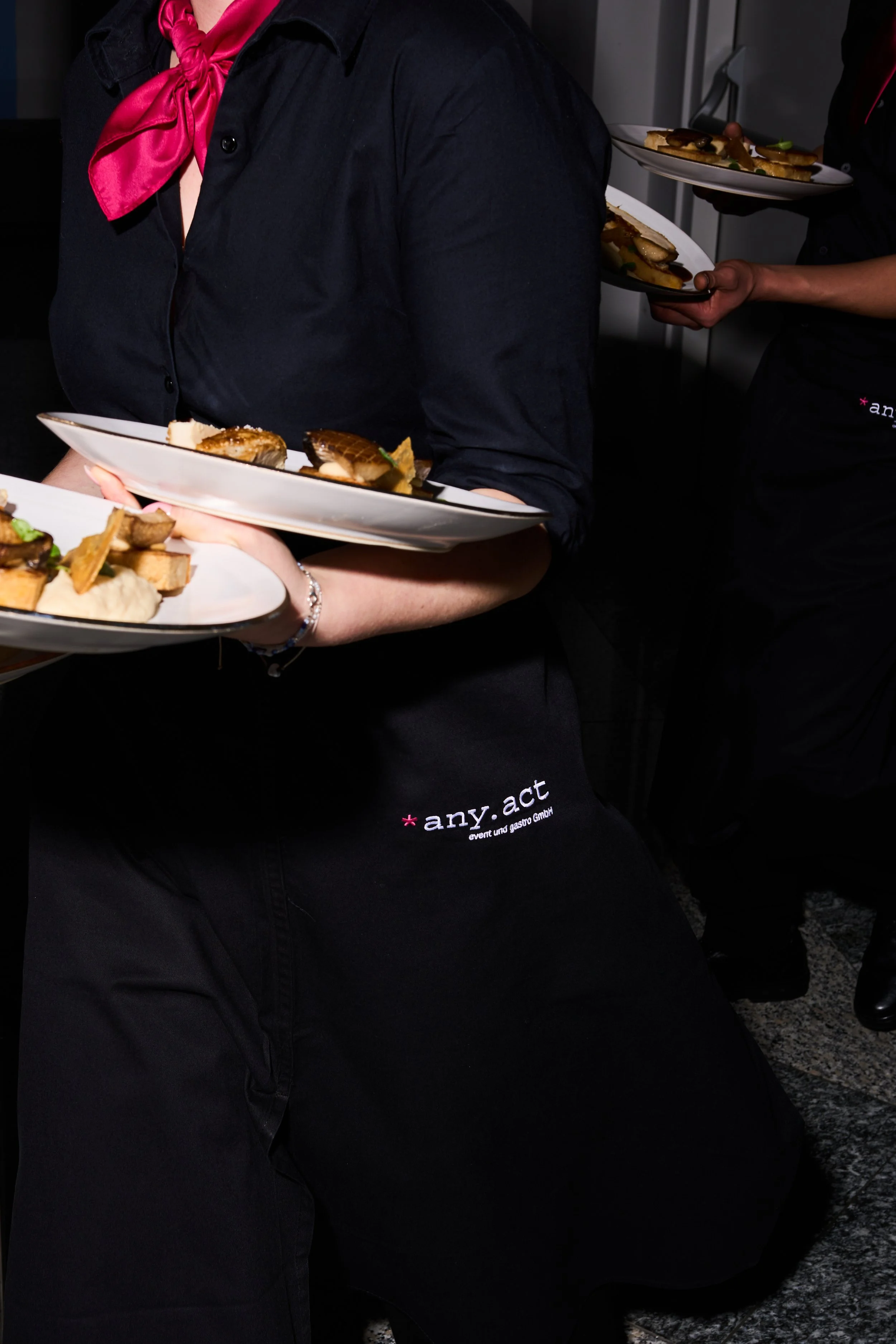 Kellnerinnen tragen schwarze uniformierte Aprons mit dem Logo 'any.act' und servieren Teller mit Essen bei einer Veranstaltung.