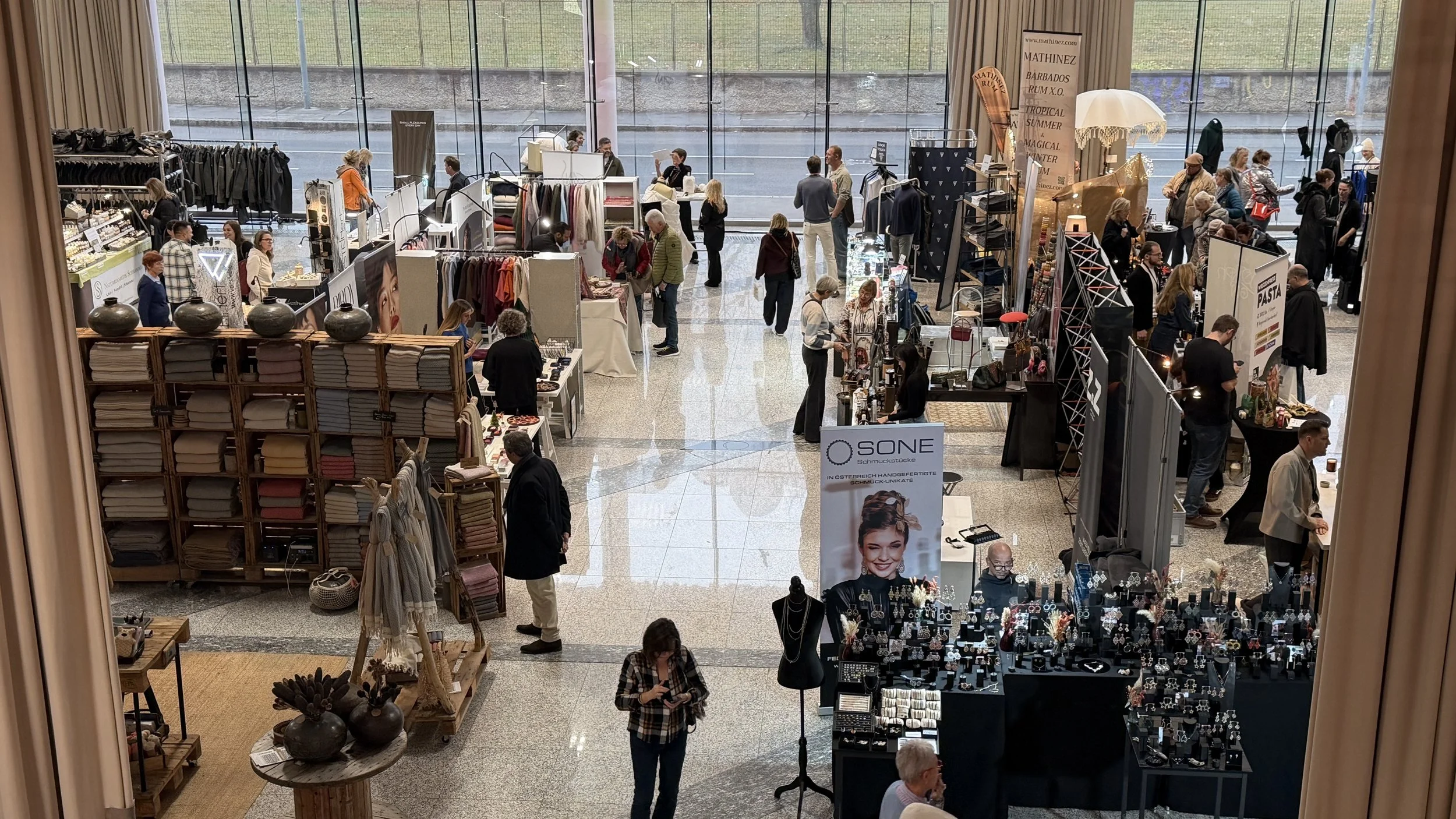 Ein belebter Indoor-Marktplatz mit verschiedenen Verkaufsständen, bei dem Menschen Kleidung, Schmuck, Kunsthandwerk und andere Waren betrachten und einkaufen, vor großen Fenstern mit Blick nach draußen.