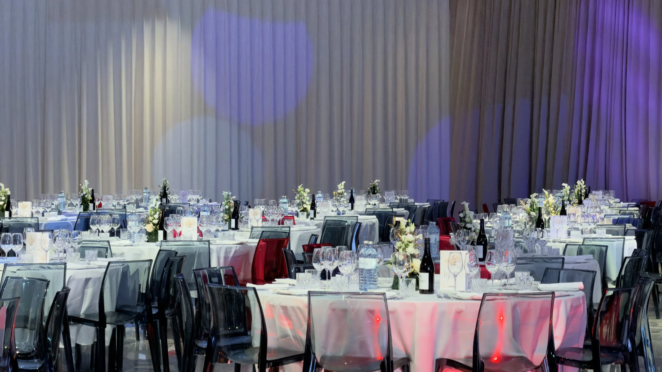 Ein festlich dekorierter Saal mit runden Tischen, weißen Tischdecken, Gläser, Flaschen, Blumenarrangements und Menükarten, beleuchtet mit buntem Licht