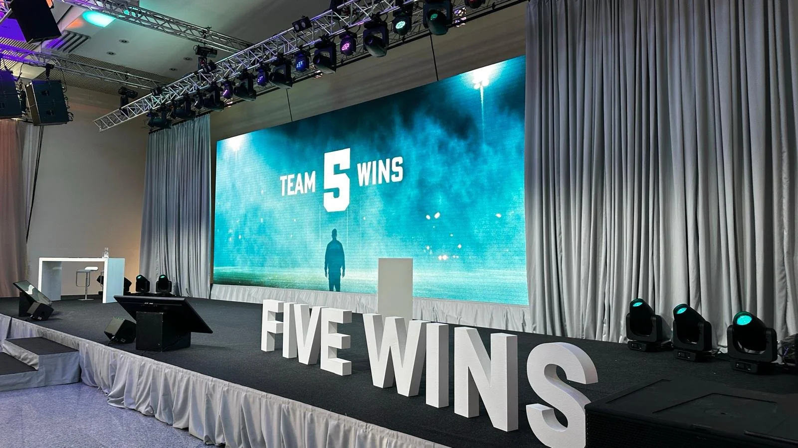 Leeres Bühne mit großem Bildschirm, der den Text 'TEAM 5 WINS' anzeigt, und große weiße Buchstaben an der Vorderkante der Bühne, die 'FIVE WINS' schreiben, vor weißen Vorhängen und Bühnenbeleuchtung.