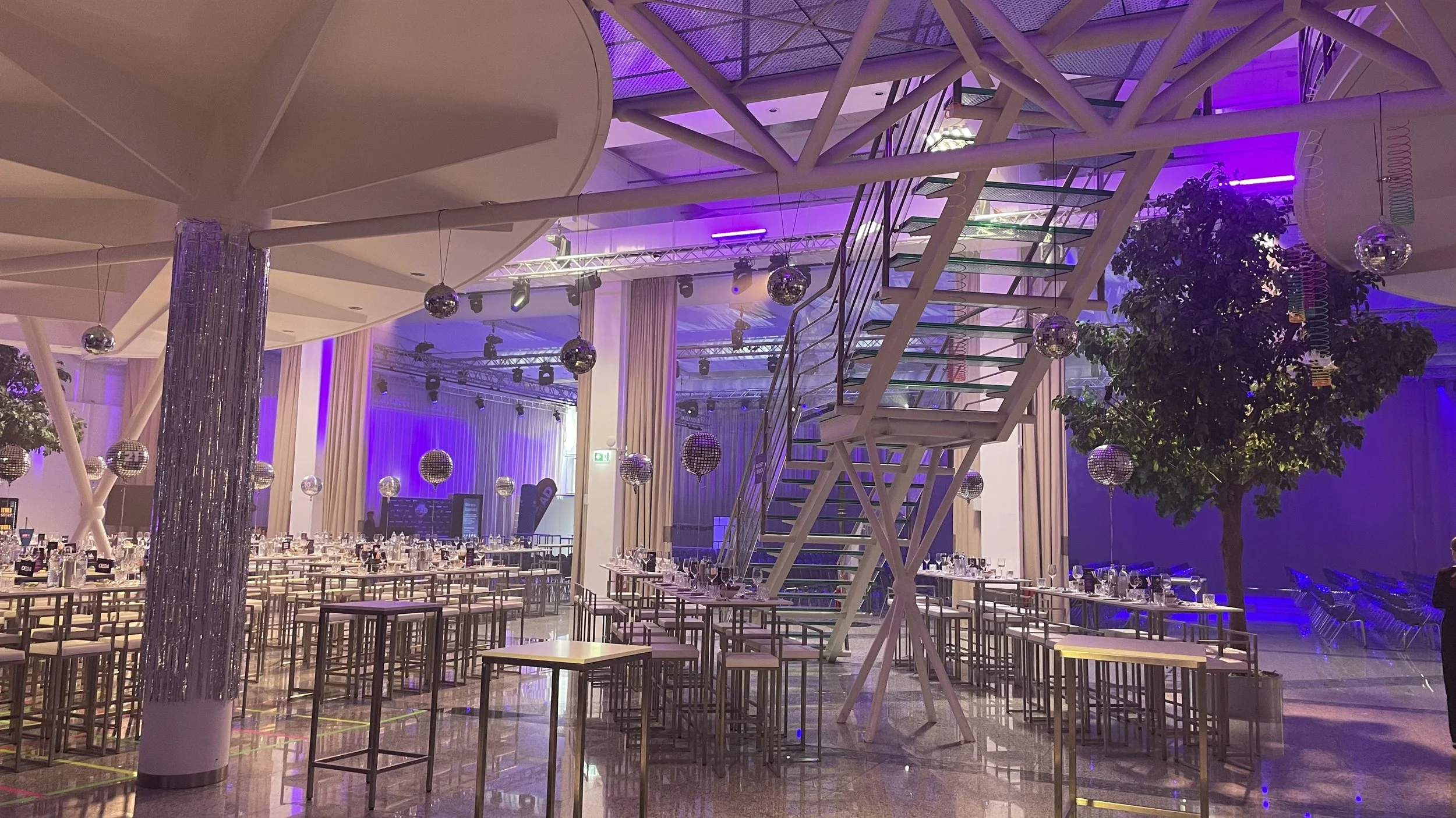 Innenraum einer festlich dekorierten Halle mit Tischen, Stühlen, Discokugeln, Lichter und einer großen Treppe.