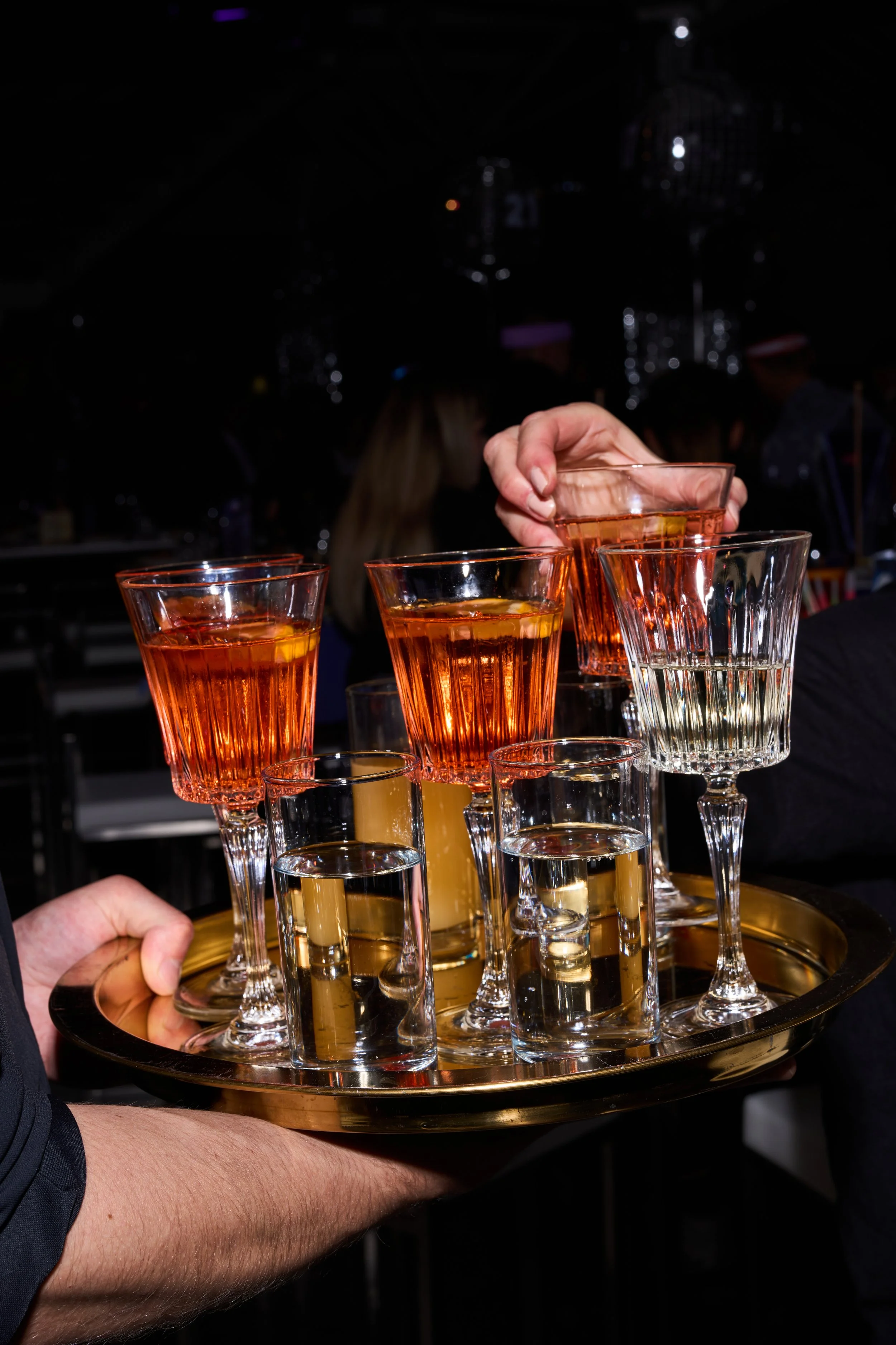 Person serviert eine Platte mit verschiedenen Gläsern, darunter Weingläser und kleine Schnapsgläser, auf einer Party.