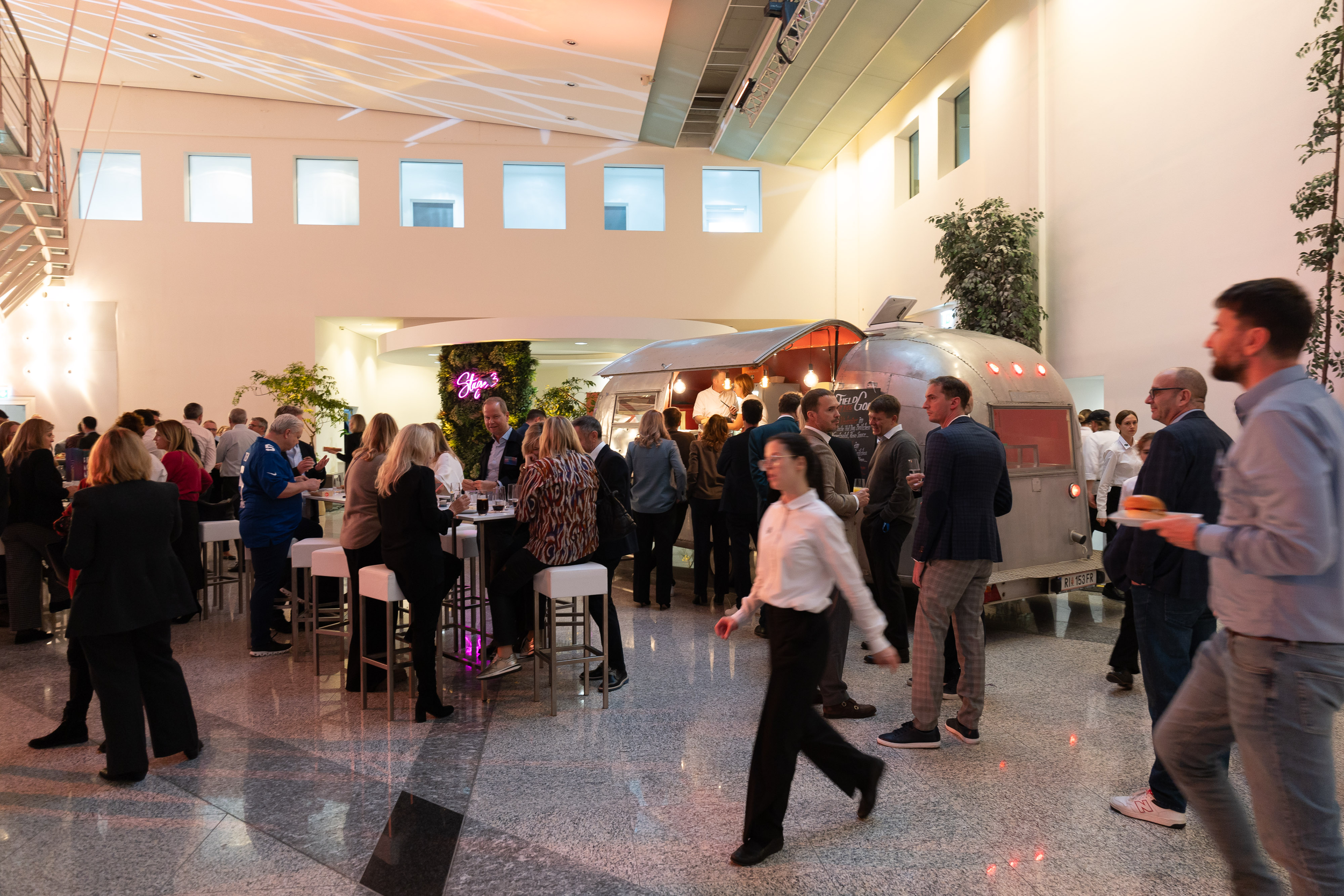 Innenansicht eines modernen Veranstaltungsortes mit einer Versammlung von Menschen, die an Steh- und Sitzbereichen essen und trinken. Im Hintergrund befindet sich eine Food-Station in einer Alttrakette. Die Raumgestaltung ist hell und offen mit hohen