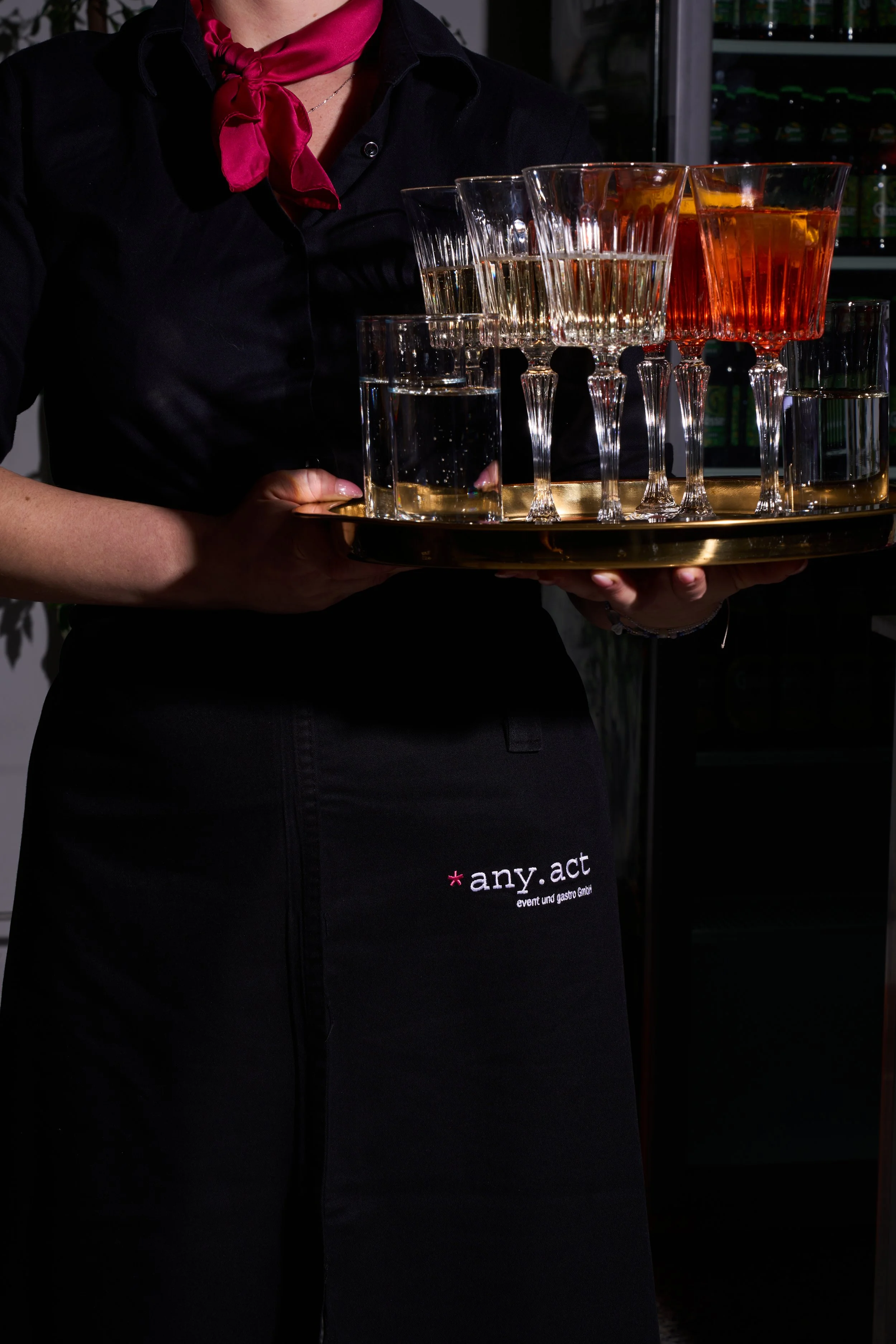 Eine Kellnerin hält einen runden goldfarbenen Tablett mit verschiedenen leeren Weingläsern, die mit unterschiedlichen Getränken gefüllt sind, in einem Bar- oder Restaurant-Umfeld.