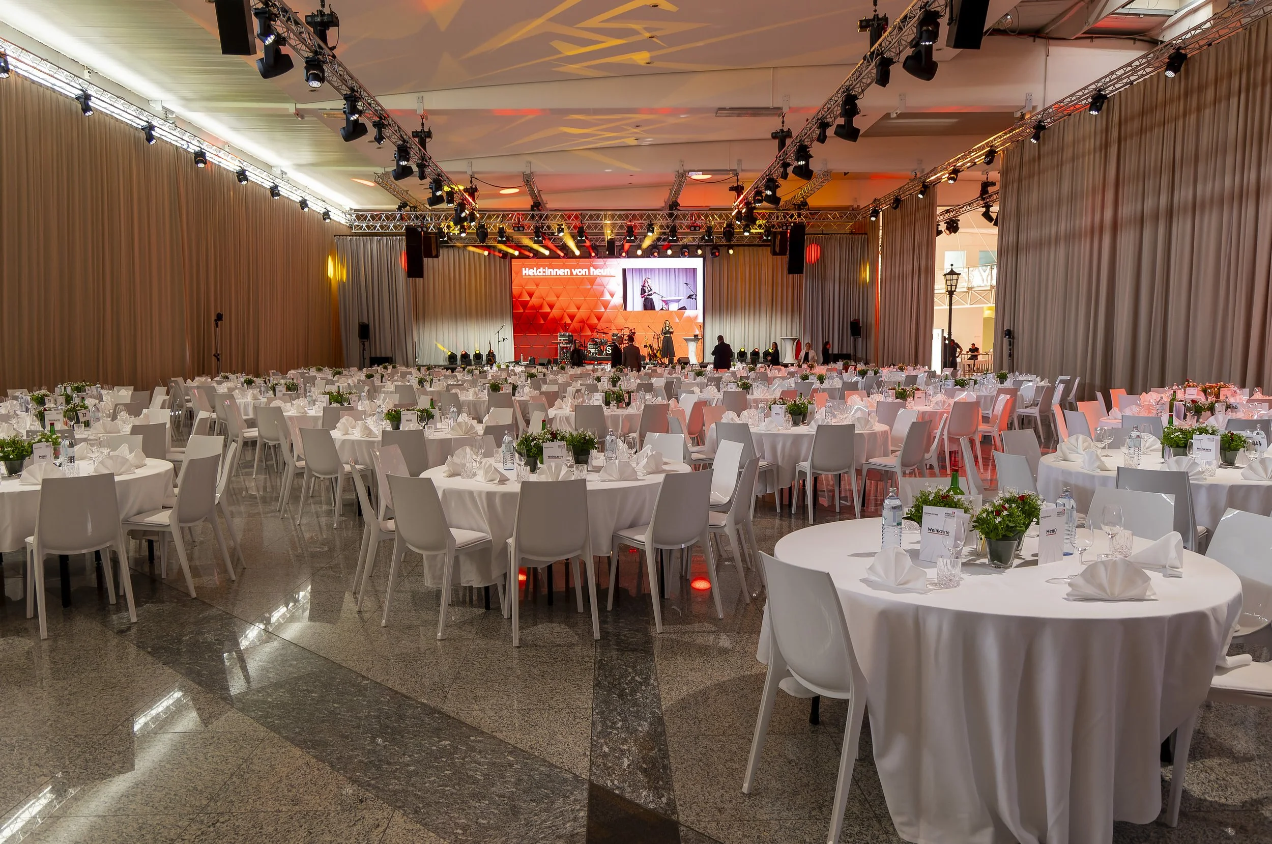Ein festlich gedeckter Saal mit runden Tischen, weißen Tischdecken, Besteck und Gläsern, mit einer Bühne im Hintergrund und Bühnenlichtern, die für eine Veranstaltung vorbereitet sind.