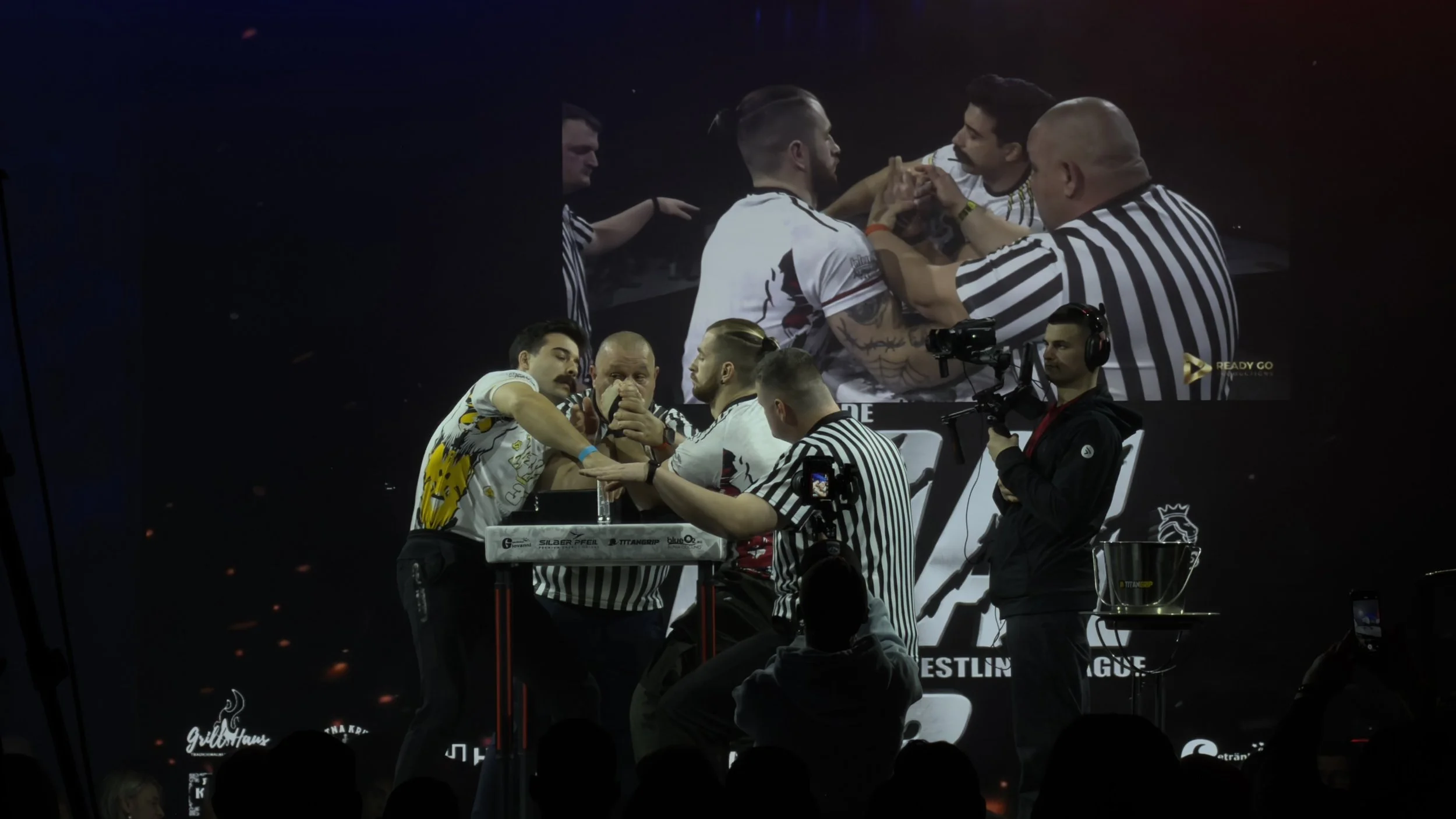Armwrestling-Wettbewerb mit zwei Männern, die um einen Tisch kämpfen, zwei Schiedsrichter überwachen das Match, im Hintergrund eine große Leinwand zeigt Nahaufnahme der beiden Kämpfer, Kameraaufnahmen aufgenommen beim Event.
