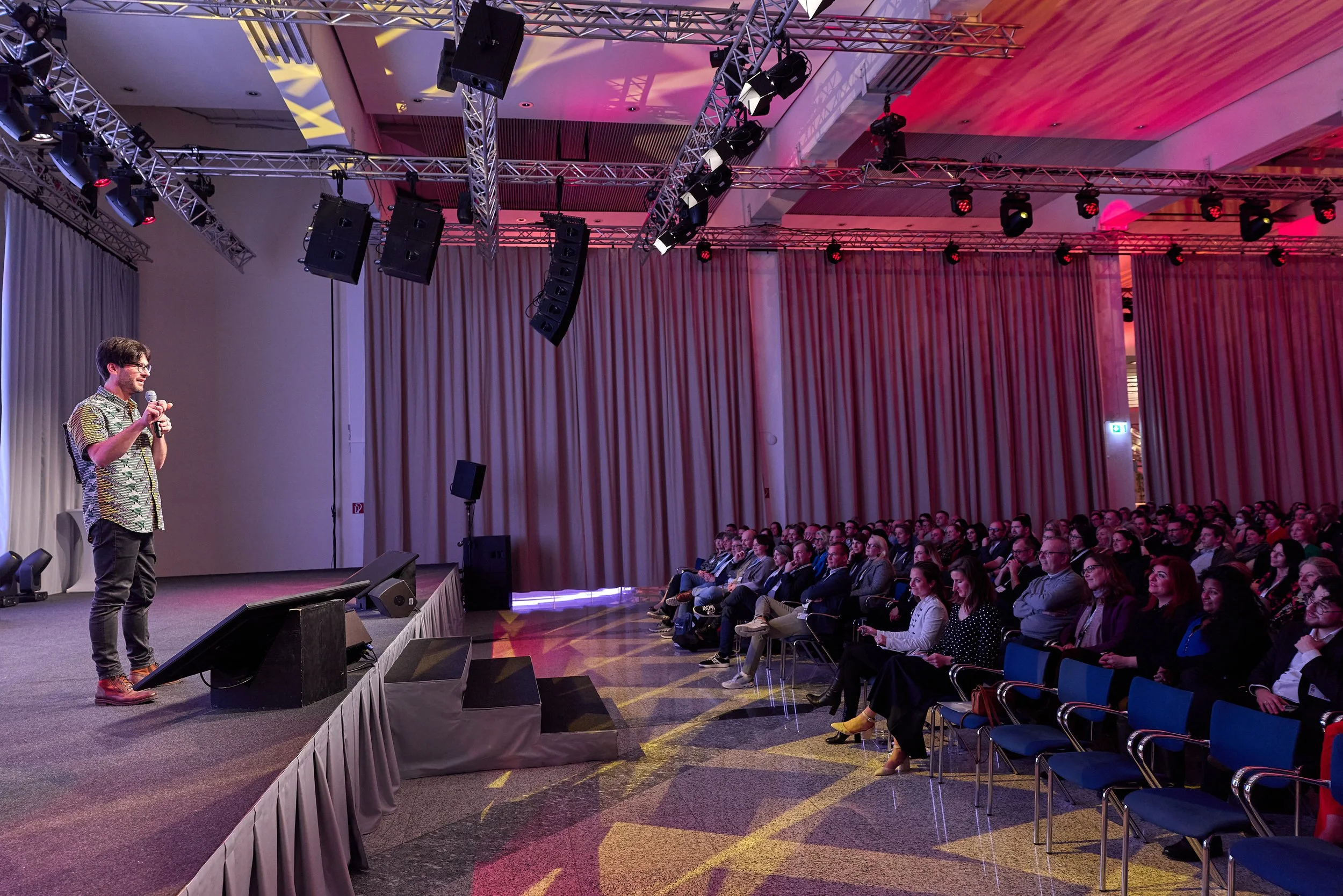 Ein Mann hält eine Rede vor einem Publikum in einem Konferenzraum mit High-End-Audio- und Lichttechnik, rote und pinke Beleuchtung, und stehendem Vorhang im Hintergrund.