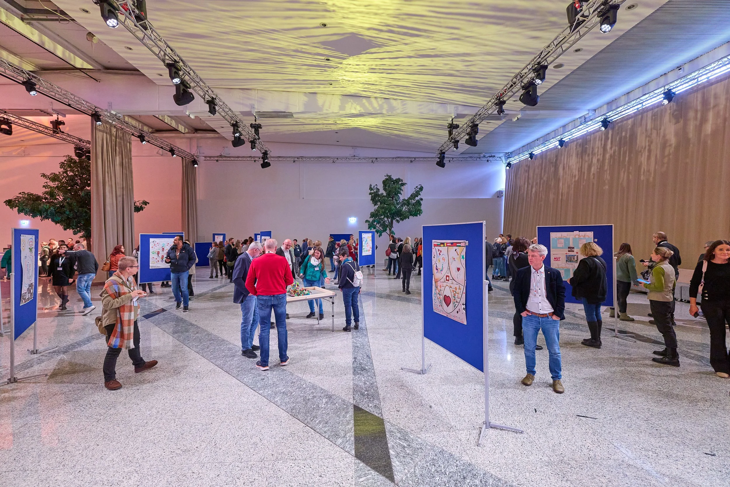 Blick in eine Kunstgalerie mit mehreren Ausstellungsbildern auf blauen Wänden. Menschen betrachten die Kunstwerke, einige stehen alleine, andere in kleinen Gruppen. Die Galerie hat eine hohe Decke mit Beleuchtung und strahlenförmige Muster auf der De