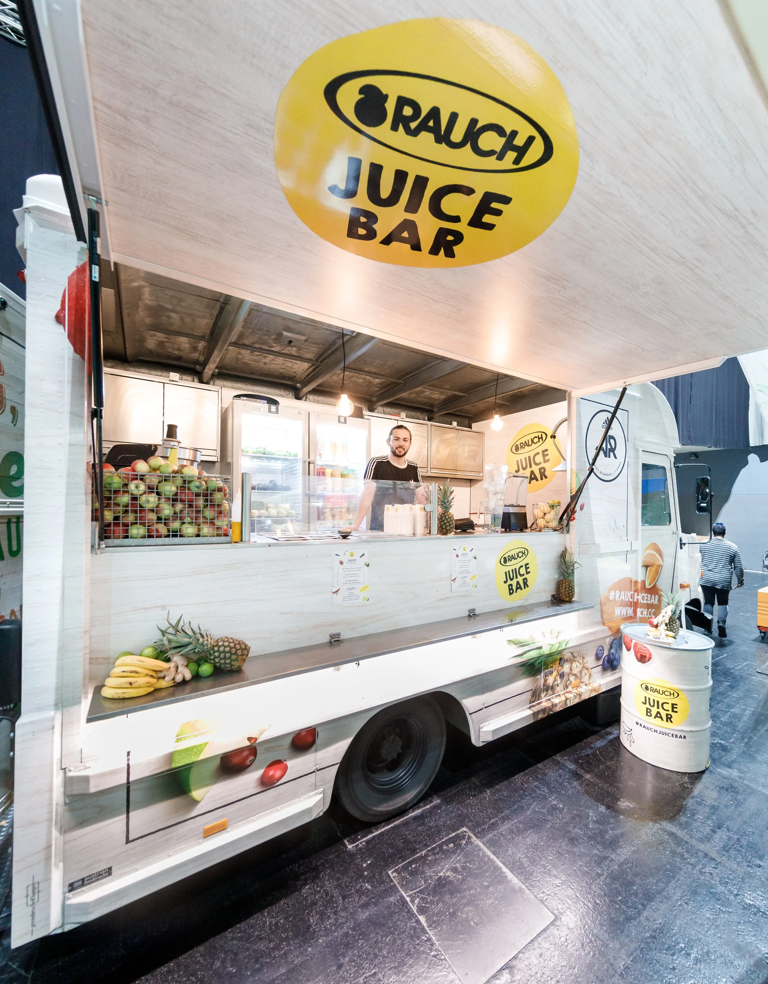 Mobiler Saftstand mit Früchten wie Bananen, Ananas, Äpfel und Limetten, Schilder mit 'Rauch Juice Bar', Verkäufer im Stand im Hintergrund.