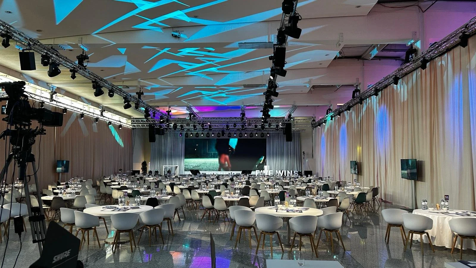 Großer Veranstaltungsraum mit runden Tischen, Stühlen, einer großen Bühne mit Bildschirm, Scheinwerfern und Vorhängen, vorbereitet für eine Firmenveranstaltung, mit mehreren Fernsehern und Wasserflaschen auf den Tischen.