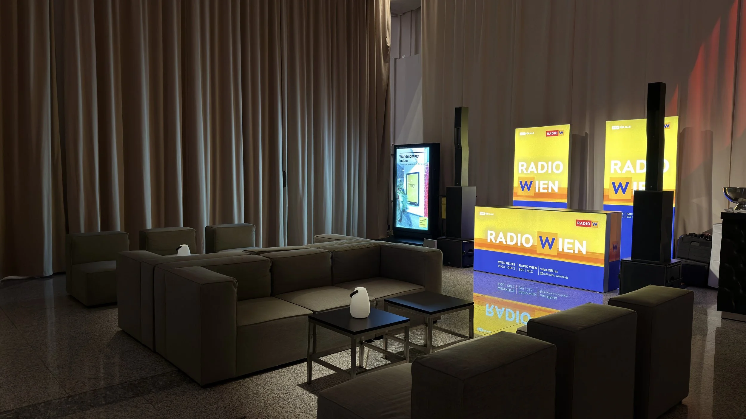 Leerer Veranstaltungsraum mit Sofa, kleinen Tischen und einem großen Bildschirm, auf dem 'Radio Wien' angezeigt wird, sowie mehrere leuchtende Werbetafeln für den Radiosender.