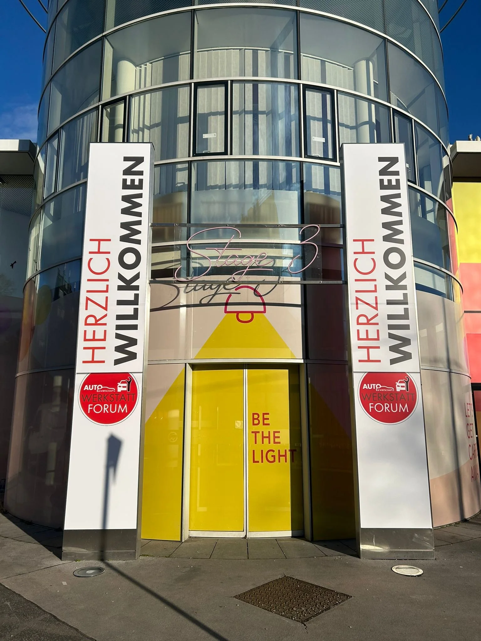Vorderansicht eines modernen Gebäudes mit großen Glasfenstern. An der Fassade befinden sich zwei vertikale Schilder mit der Aufschrift "HERZLICH WILLKOMMEN". Über dem Eingang ist eine neonbeleuchtete Signatur mit dem Wort "Stage 3". Über der Tür steh