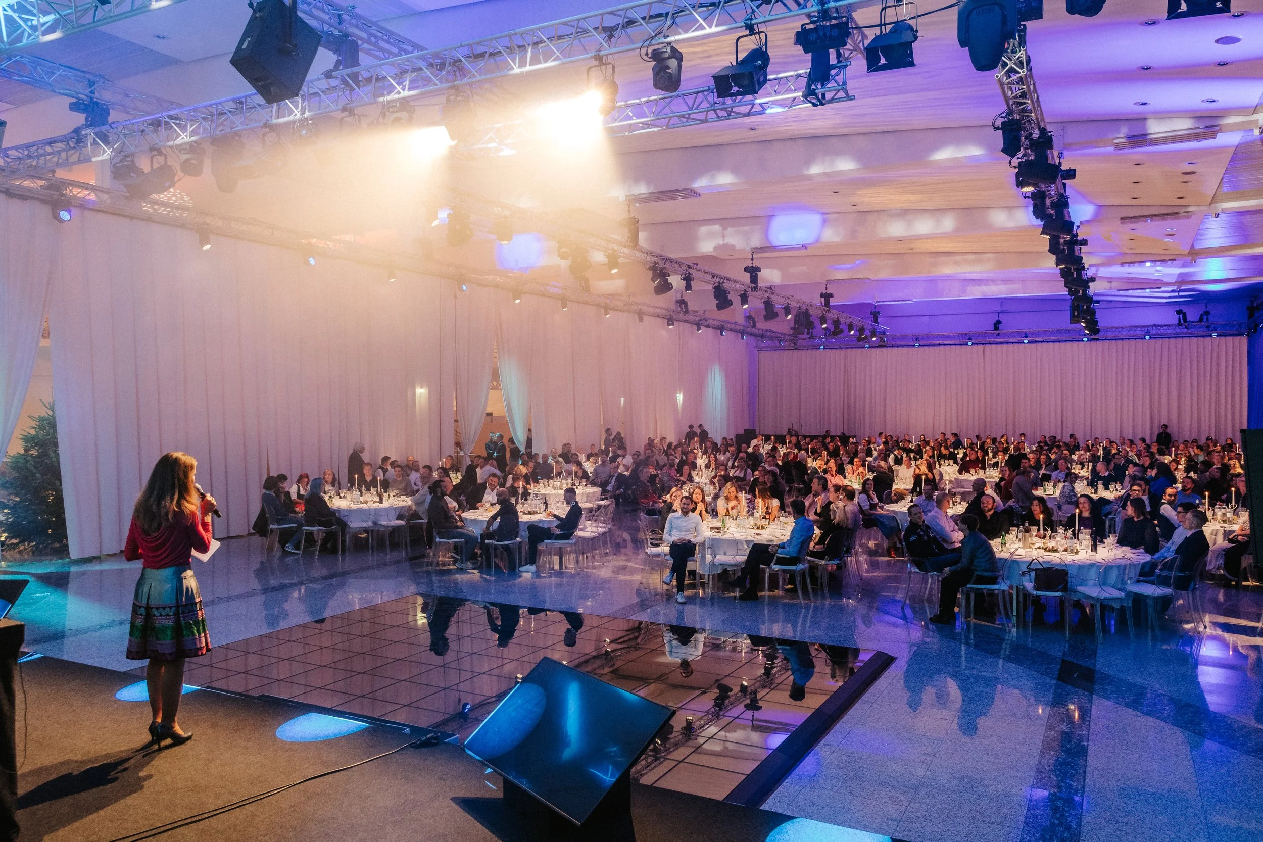 Nousiert und lichterfüllter Veranstaltungsraum mit vielen Tischen und Gästen, die einem Redner auf der Bühne zuhören.