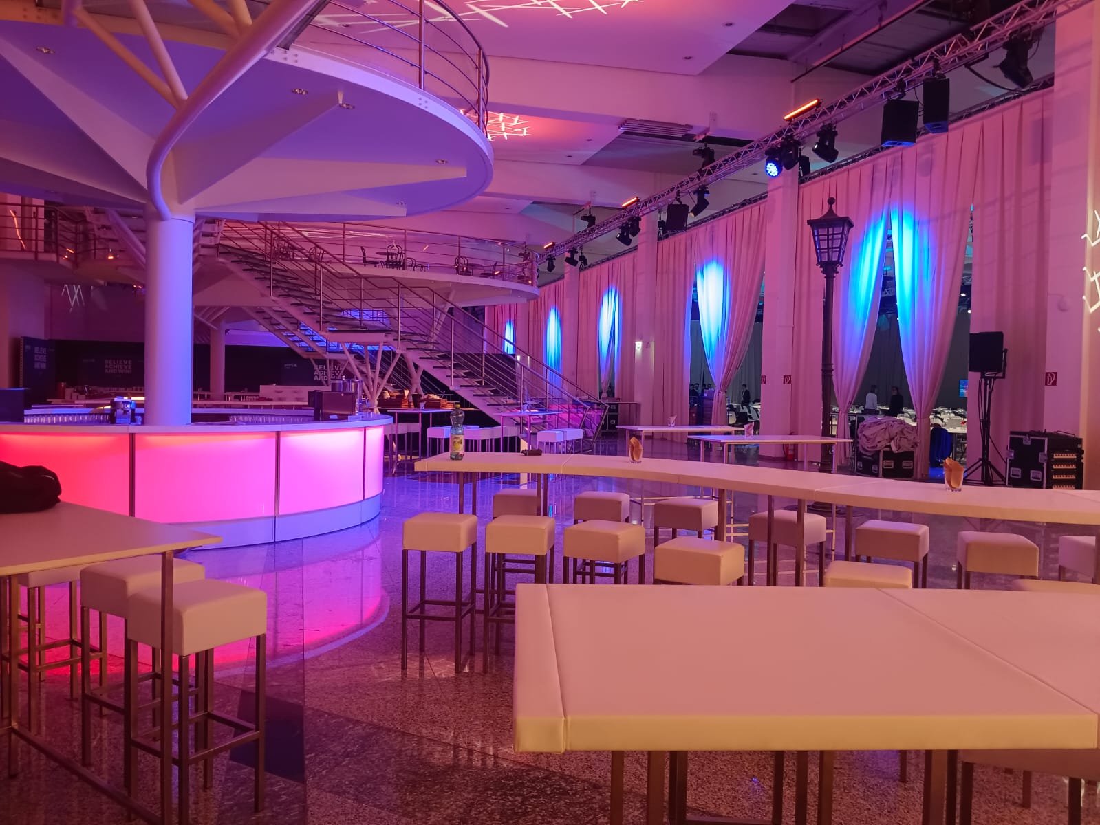 Leeres Veranstaltungsraum mit Bar, Tischen, Stühlen, Beleuchtung in Blau- und Pinktönen, Vorhängen und Bühne mit Lichttechnik.