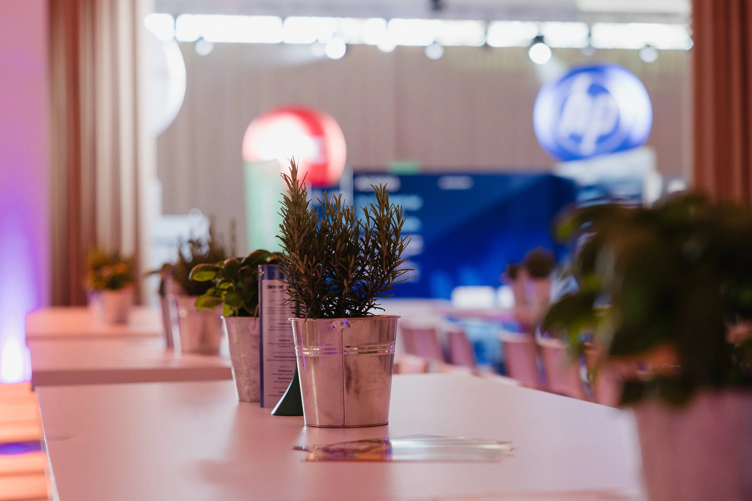 Innenraum eines Veranstaltungsraums mit Blumenkübeln auf weißen Tischen, im Hintergrund bunte Lichter und große Firmenlogos.