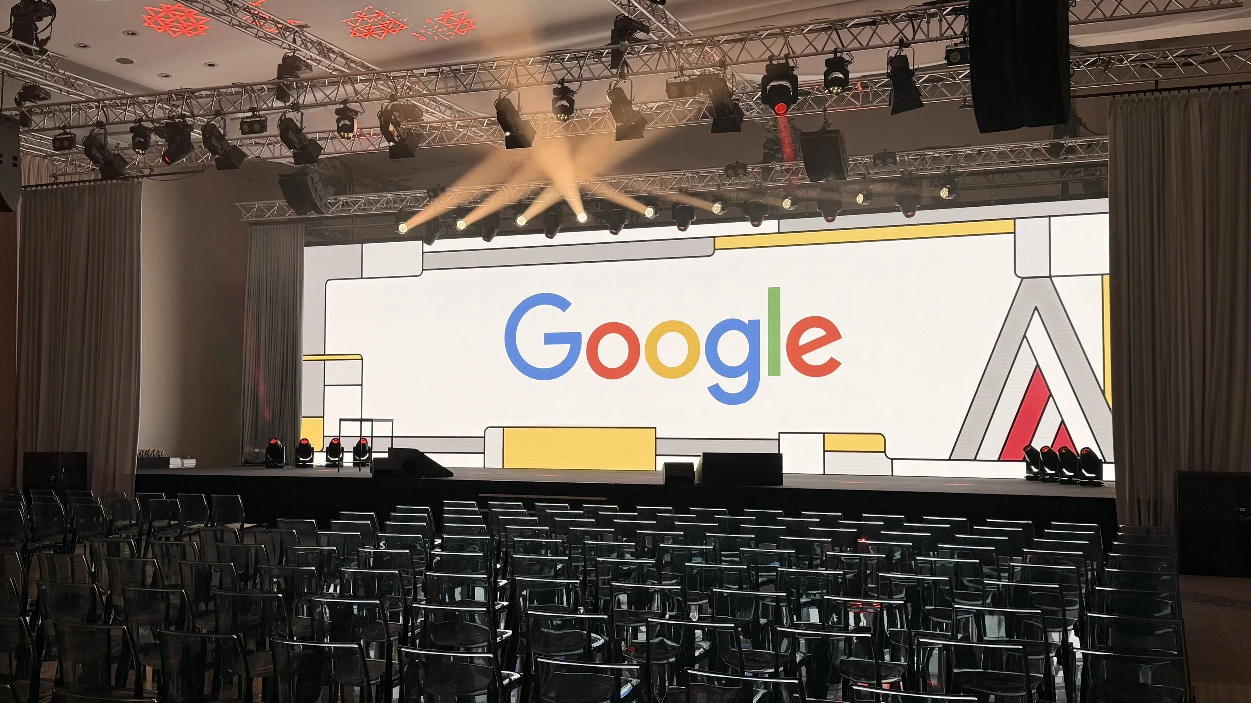 Leeres Veranstaltungszimmer mit einer großen Bühne, einem großen Bildschirm mit Google-Logo, mehrere Scheinwerfer und Stühle, die auf die Bühne ausgerichtet sind.