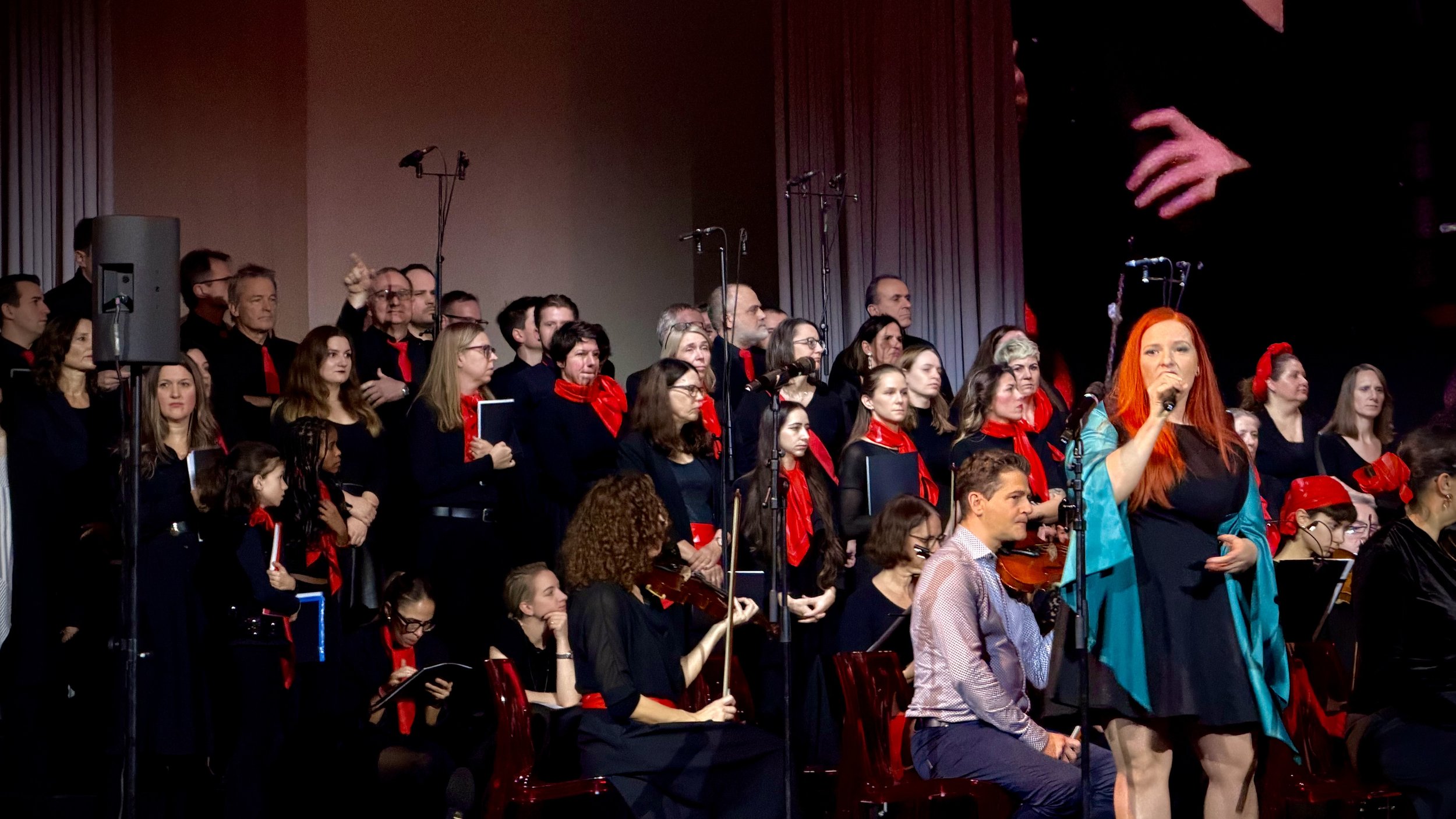Chor auf einer Bühne, viele Sänger in schwarzen Kleidern und Anzügen mit roten Halstüchern, eine Frau mit rotem Haar singt in Mikrofon und trägt einen schwarzen Rock und eine blaue Jacke.