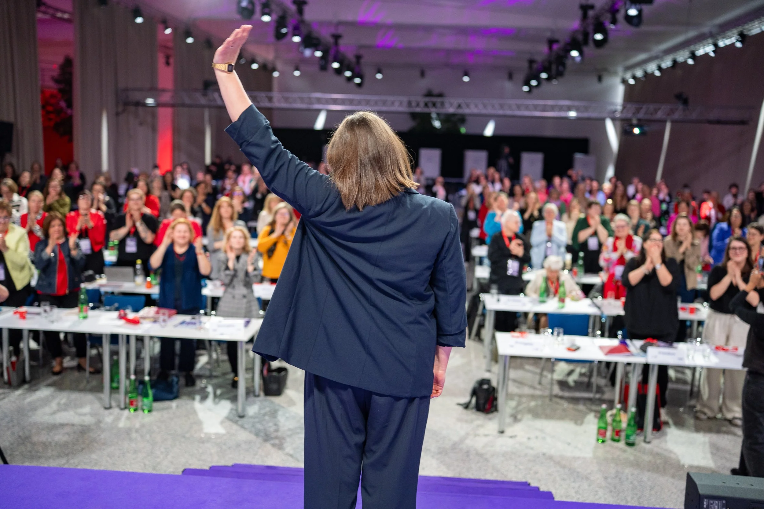 Eine Frau steht auf einer Bühne vor einem Publikum und winkt mit der Hand. Das Publikum besteht aus Männern und Frauen, die applaudieren und lächeln. Es ist eine Konferenz oder ein Seminar in einem großen Saal.