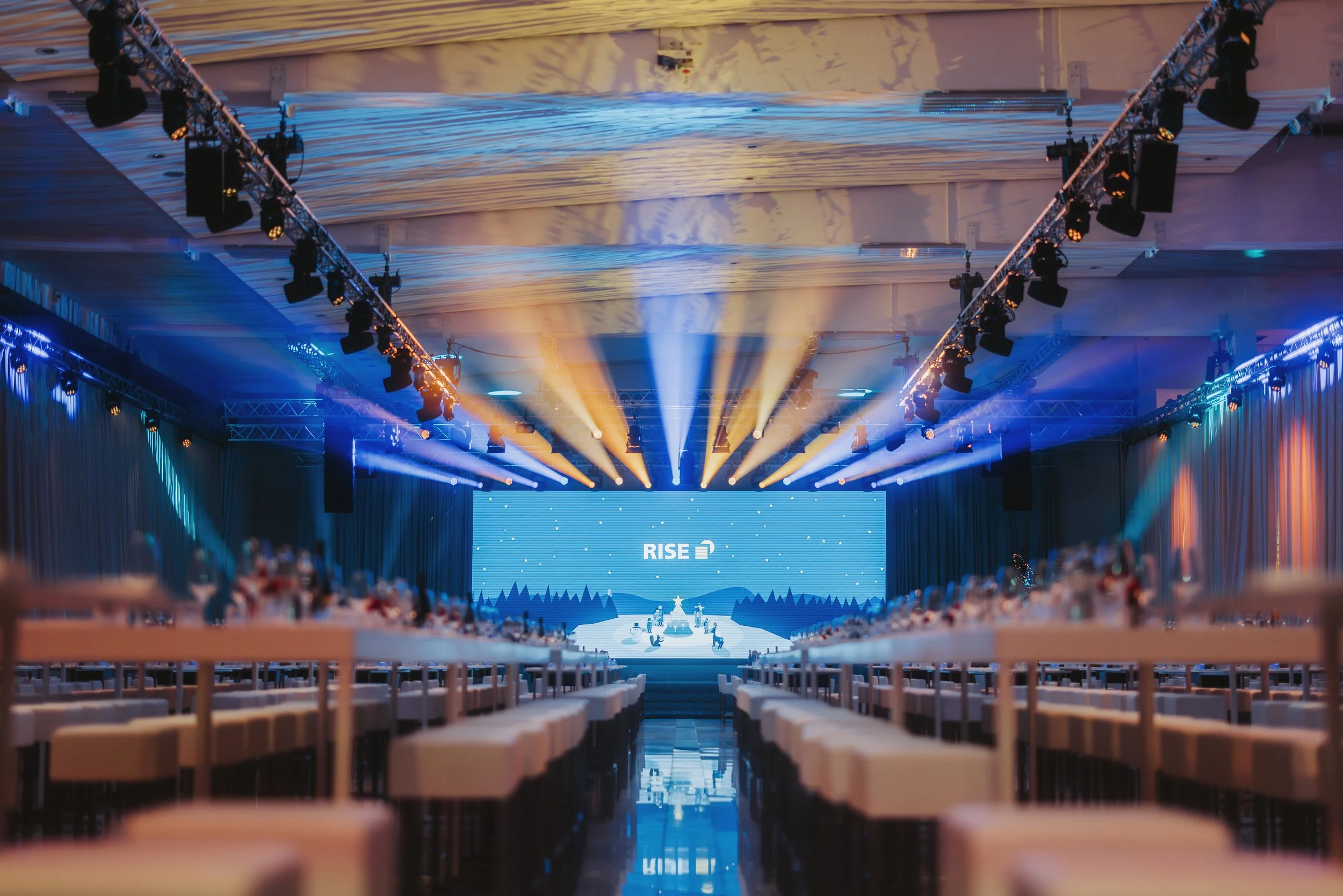Ein großer Veranstaltungsraum mit einer Bühne, blauen und warmen gelben Scheinwerfern, Vorhängen an den Seiten und Tischen mit Stühlen, die für eine Konferenz oder Gala vorbereitet sind. Auf der Bühne ist ein großer Bildschirm mit der Aufschrift 'RIS