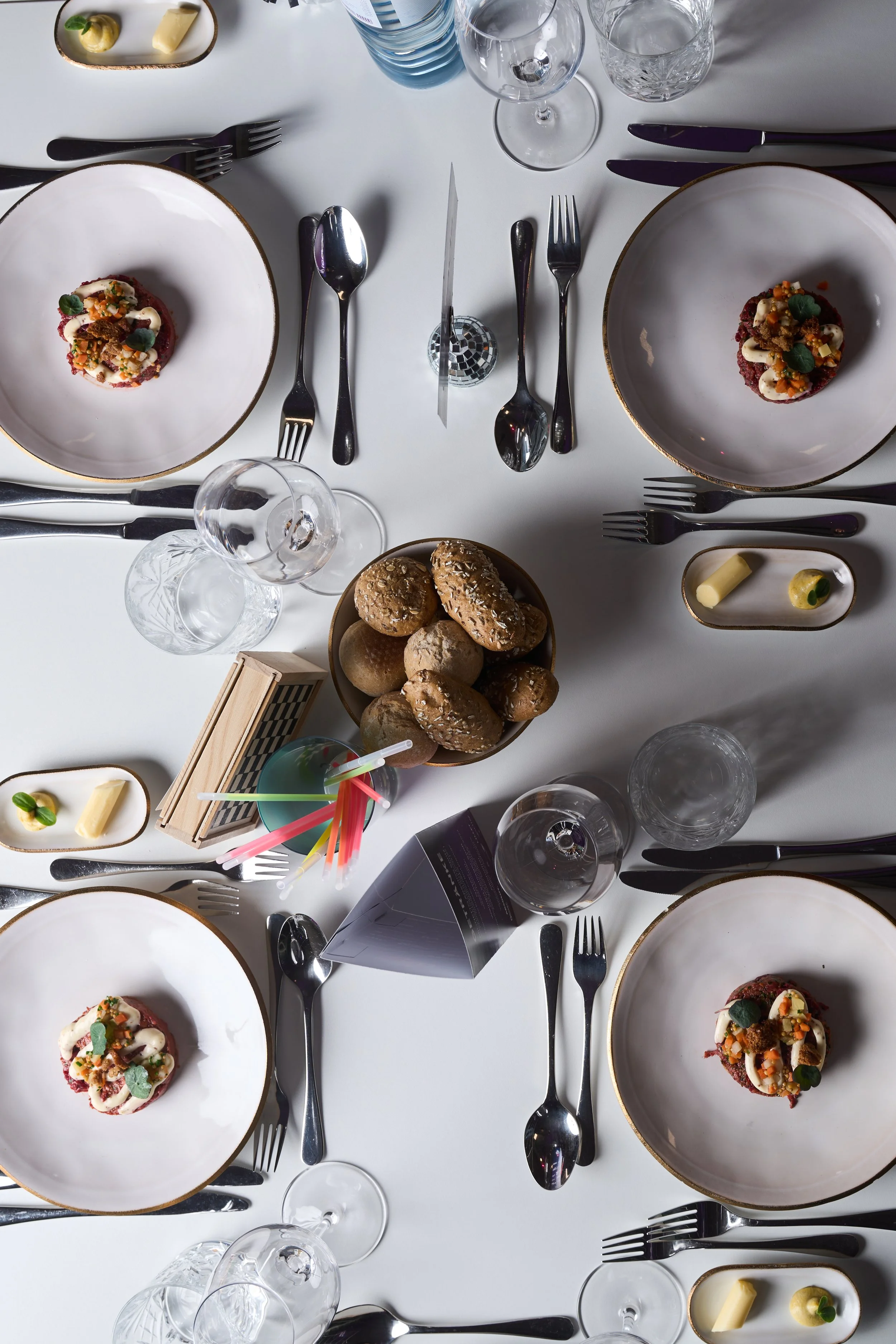 Tisch mit vier Tellern, voll mit kleinen Speisen, mehreren Gläsern, Besteck, sowie einem Korb mit Brötchen und weiteren Dekorationen.