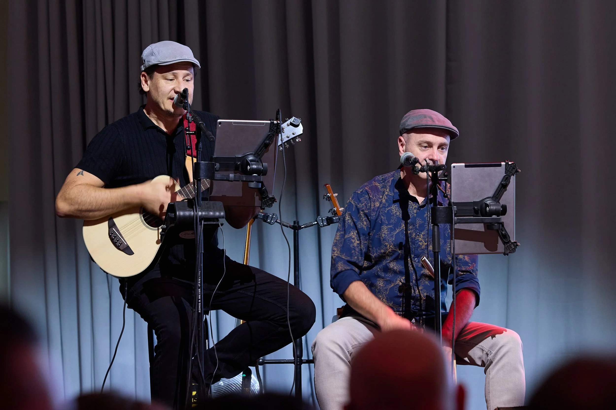 Zwei Männer spielen auf einer Bühne, einer mit Gitarre, beide tragen Hüte, vor einem Vorhang.