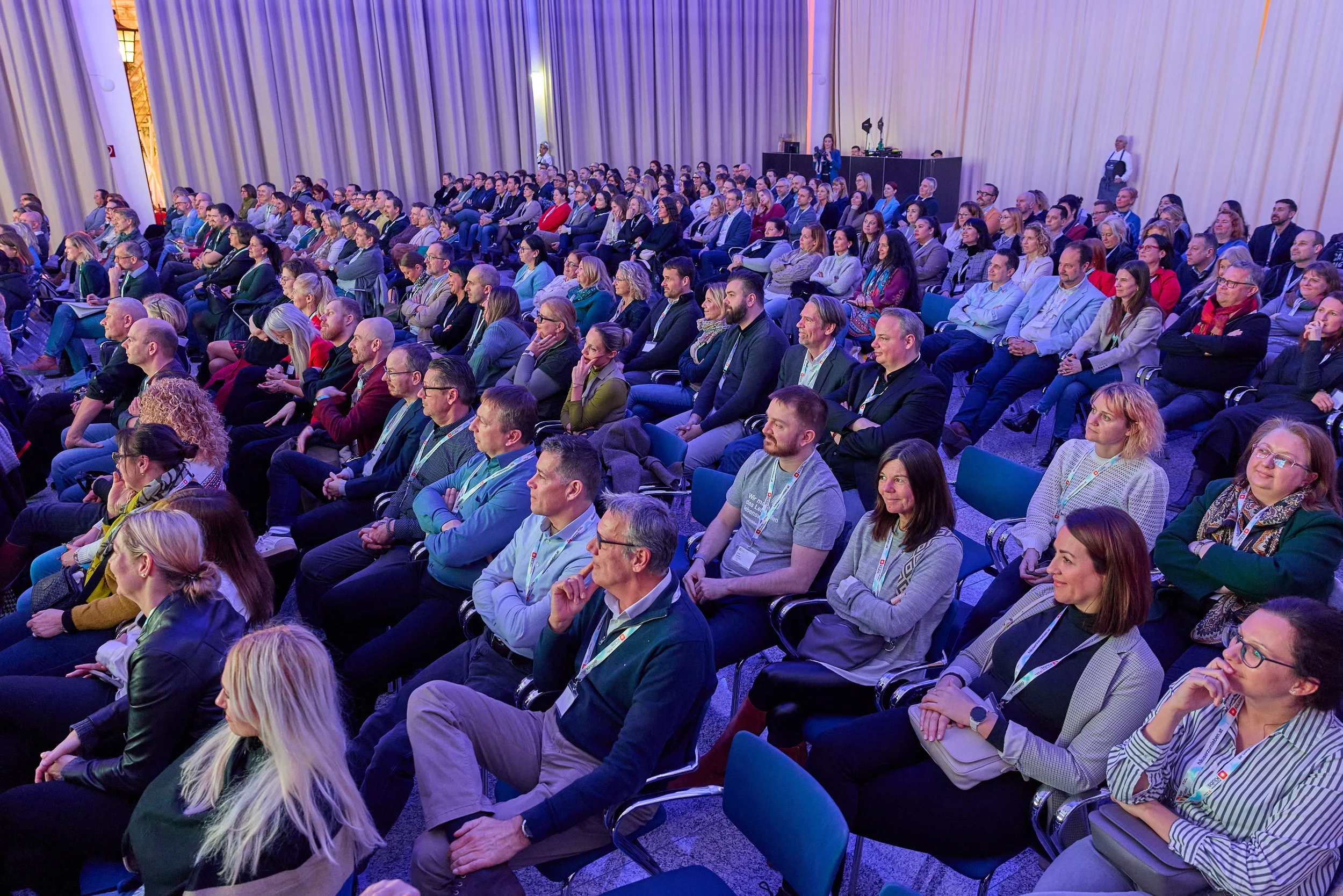 Konferenzraum voller gehörter Menschen, die einem Vortrag oder einer Präsentation lauschen, mit einer Bühne im Hintergrund.