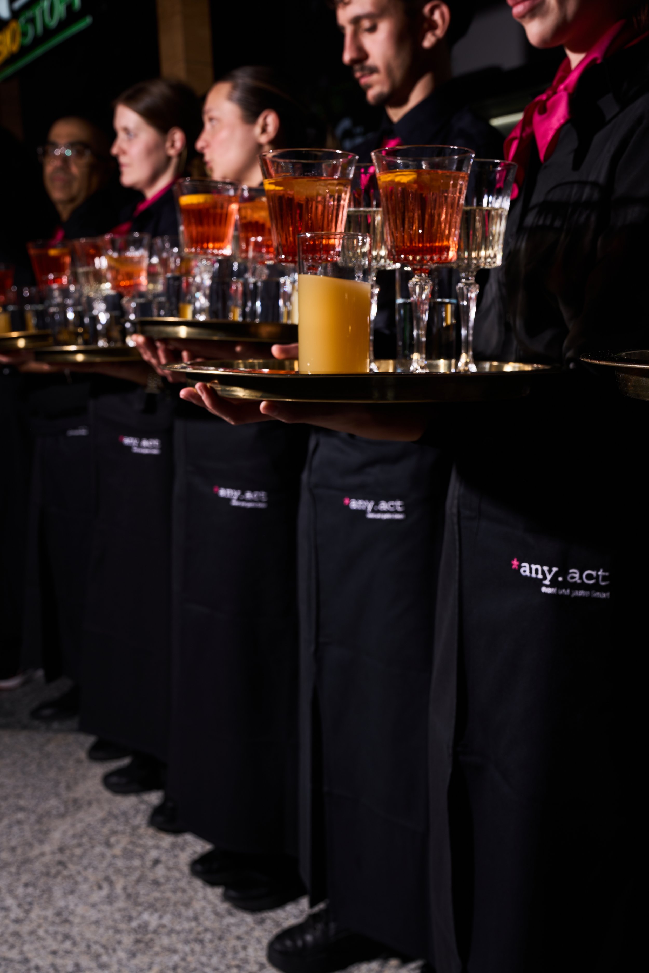 Mehrere Kellner in schwarzen Schürzen, die Getränke auf goldenen Tabletts servieren, während sie in einer Reihe stehen, in einem dunklen Raum