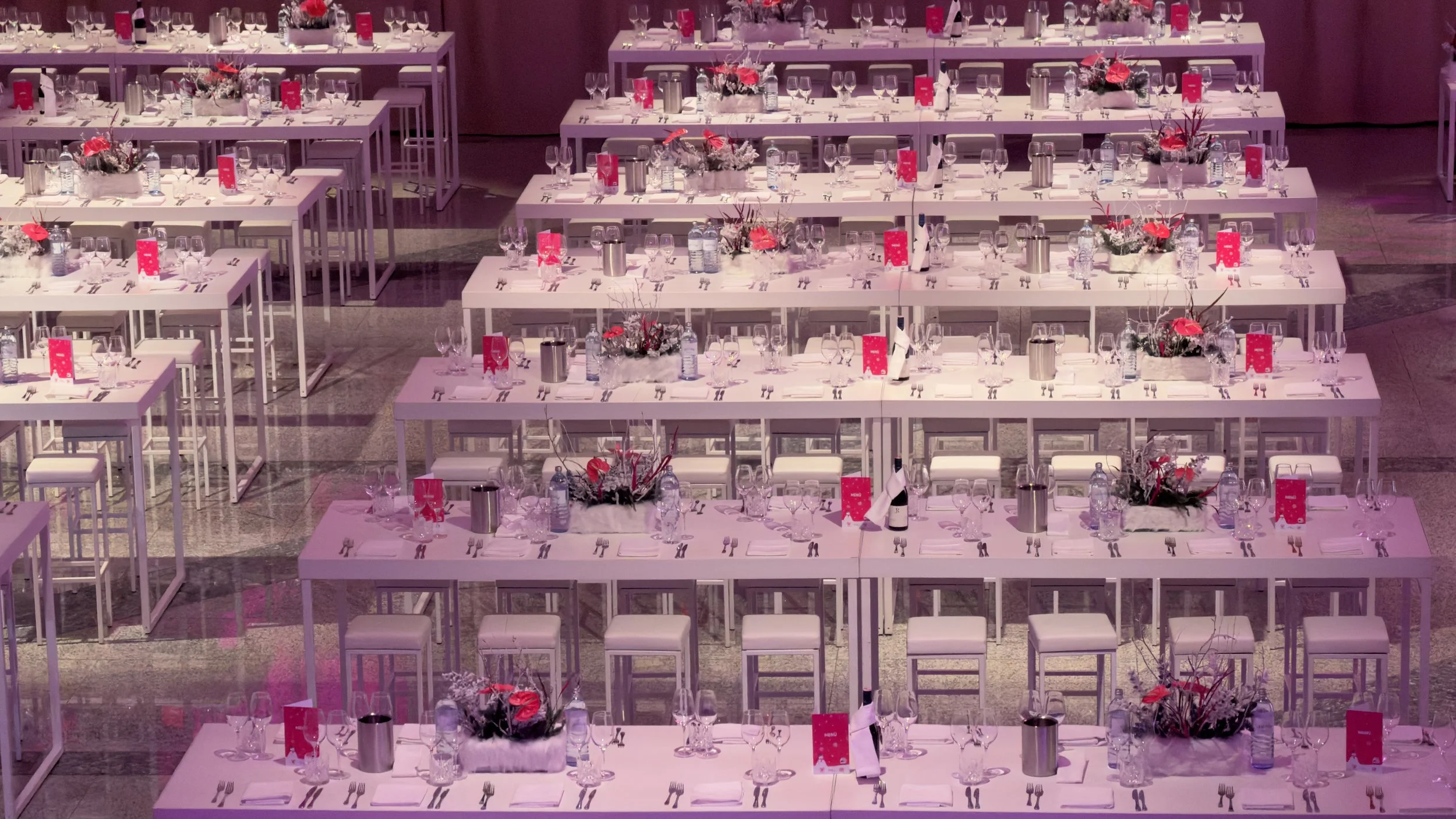 Mehrere lange, weiße Tische, die für eine Veranstaltung gedeckt sind. Die Tische sind mit Gläsern, Besteck, weißen Servietten, Blumenarrangements und roten Menükarten dekoriert.