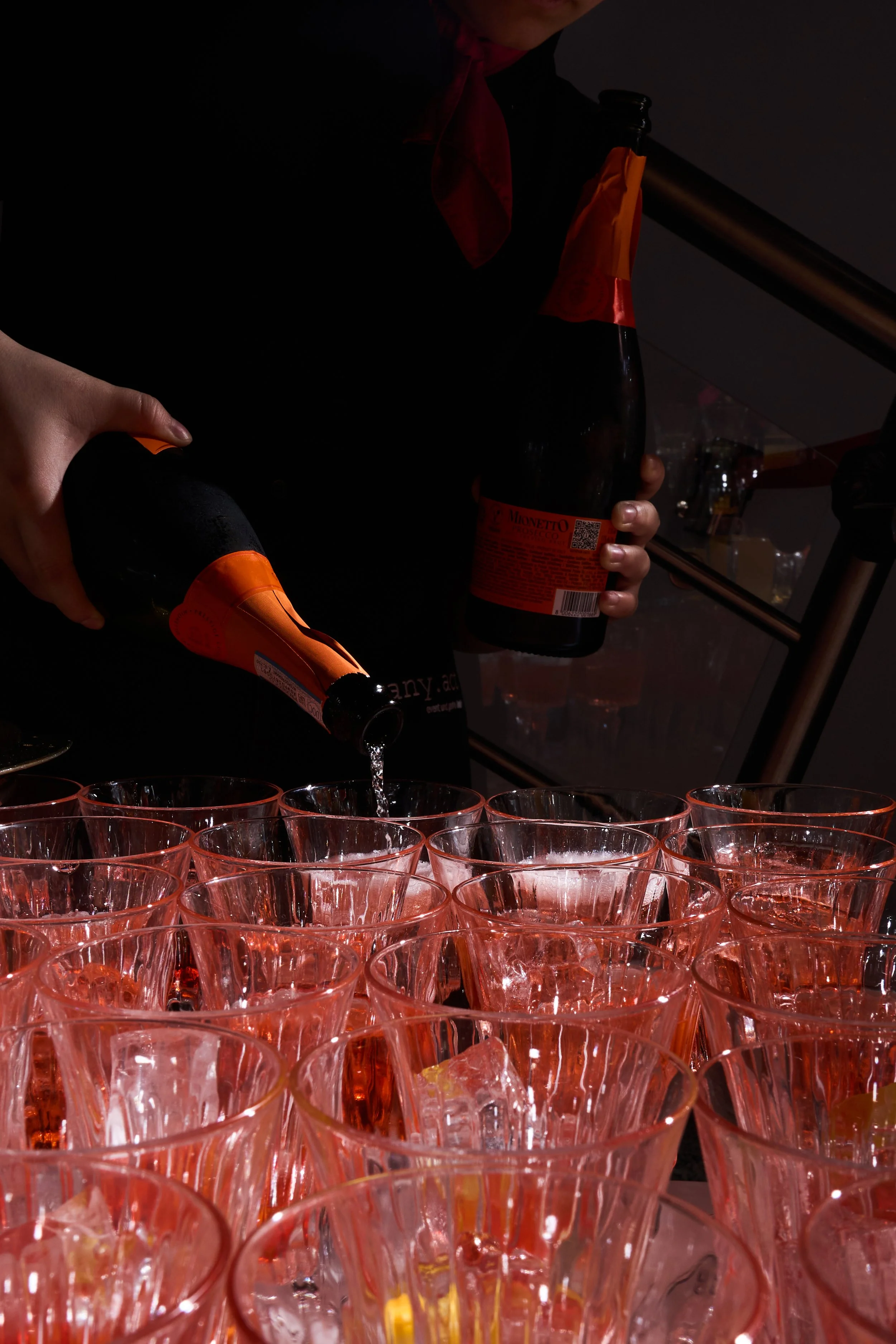 Person in dunklem Outfit und rotem Halstuch schenkt Sekt in Glas mit Eis bei einer Party