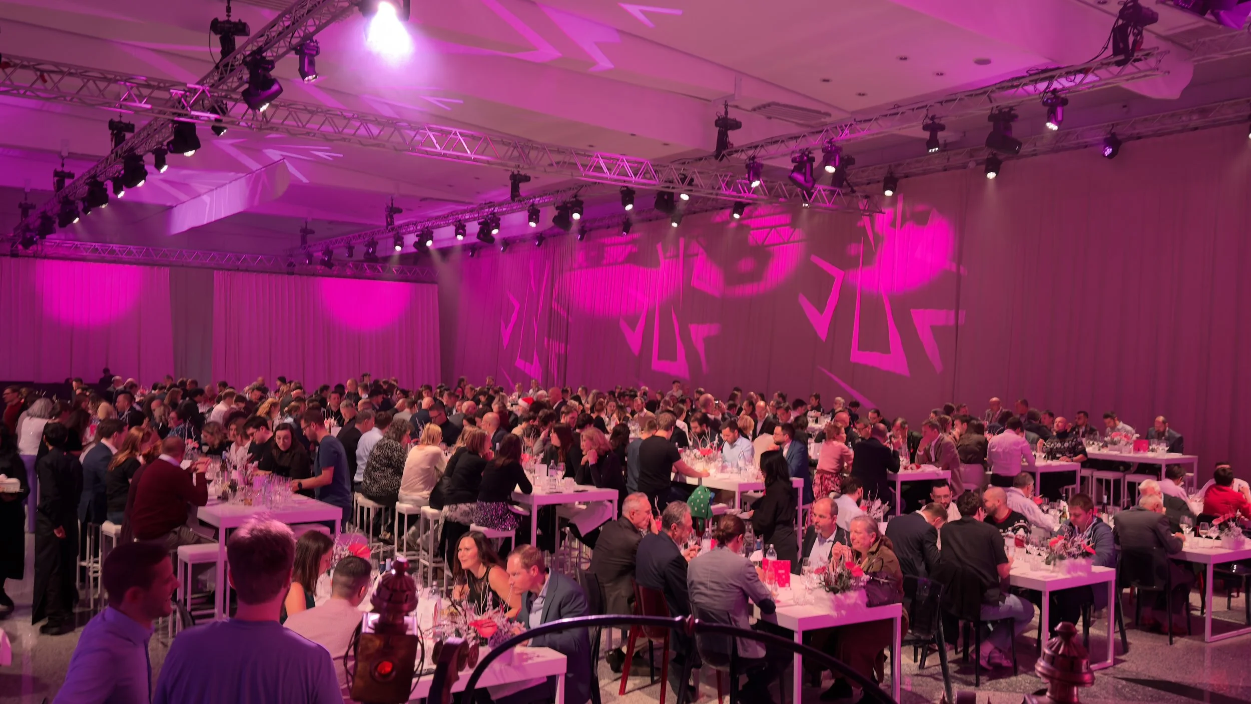 Große Gruppe von Menschen bei einem formellen Abendessen in einem Hotel oder Veranstaltungsraum mit pinker Beleuchtung und dekorativen Lichteffekten an den Wänden.