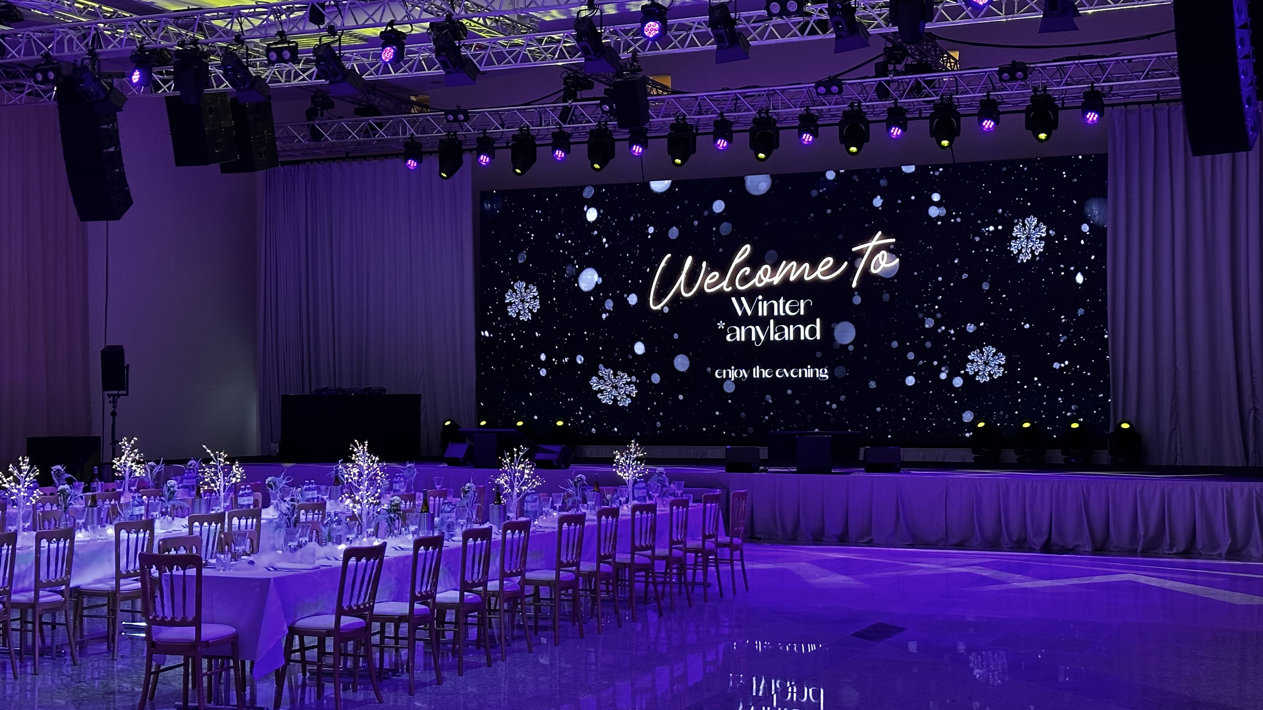 Blick auf eine festliche Winterveranstaltung mit einem großen Bildschirm, der 'Welcome to Winter anyland' und 'enjoy the evening' anzeigt. Der Raum ist mit eleganten Tischen, weißen Stühlen, winterlichen Dekorationen und leuchtenden Winterlichtern de