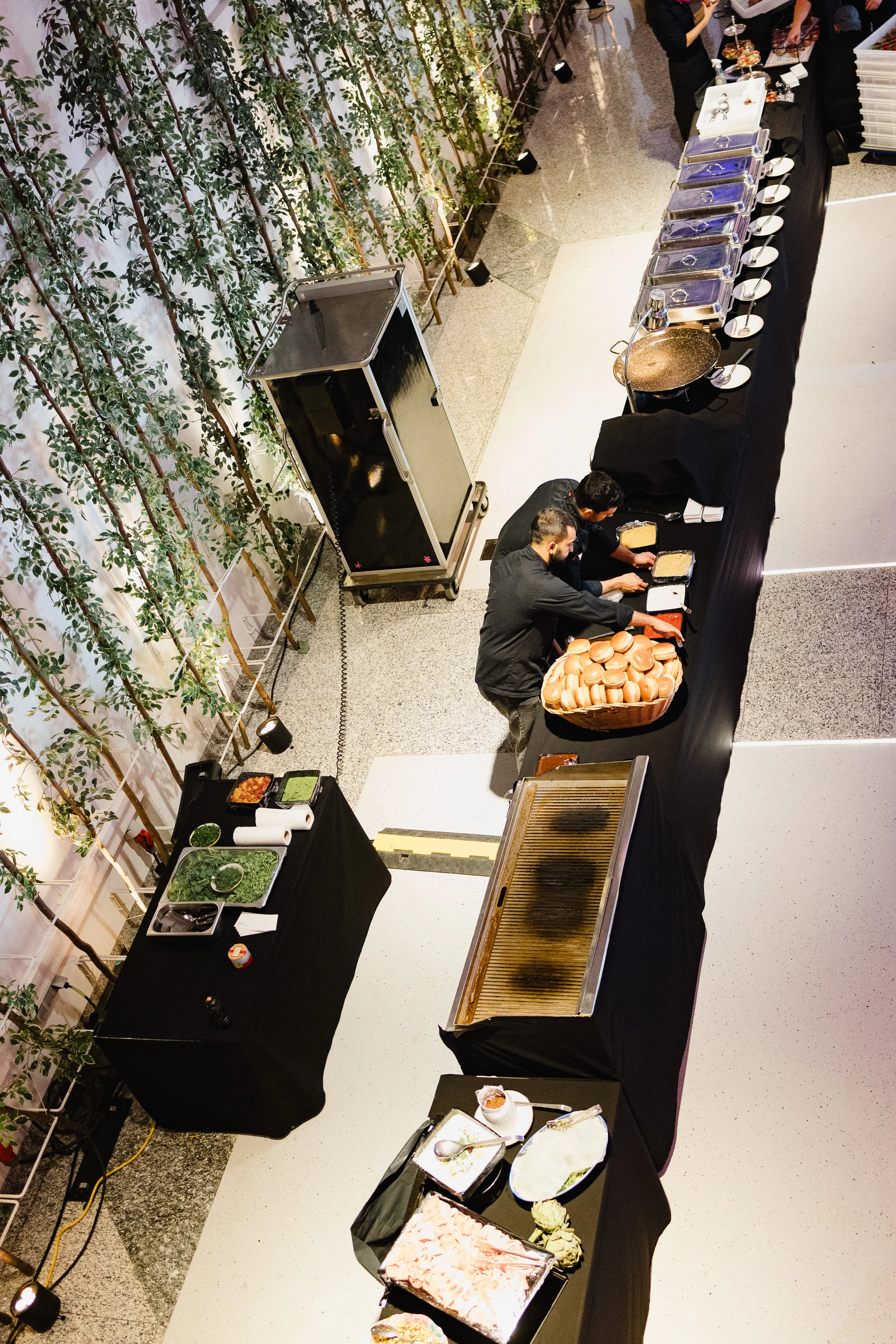Blick auf ein Buffet bei einer Veranstaltung, mit mehreren Köchen, die Sandwiches zubereiten, und verschiedenen Speisen auf Tischen, darunter Hamburger Brötchen, Salate und Desserts.