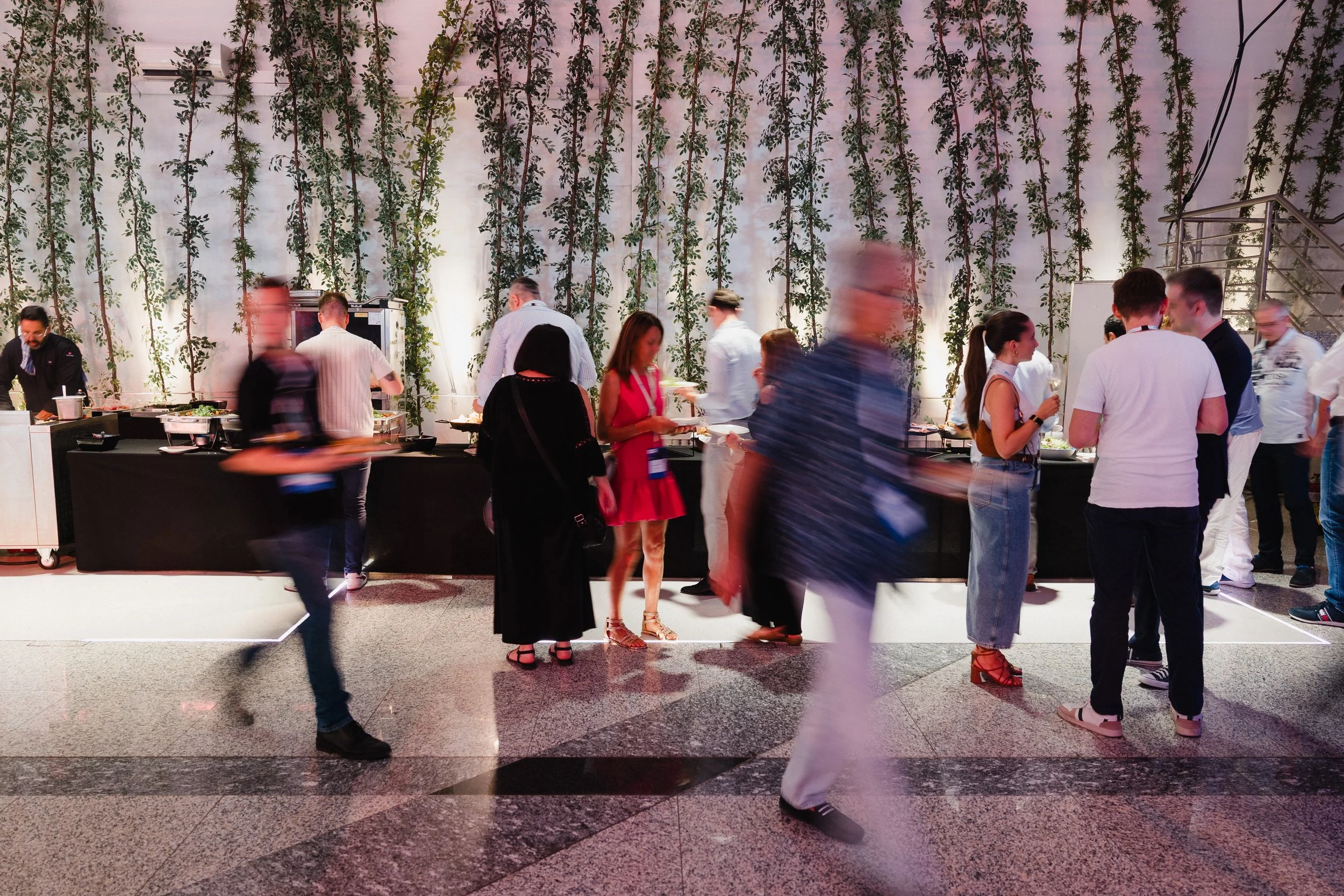 Menschen bei einem Buffet mit einer weißen Wand im Hintergrund, die mit grünen Pflanzen bedeckt ist.