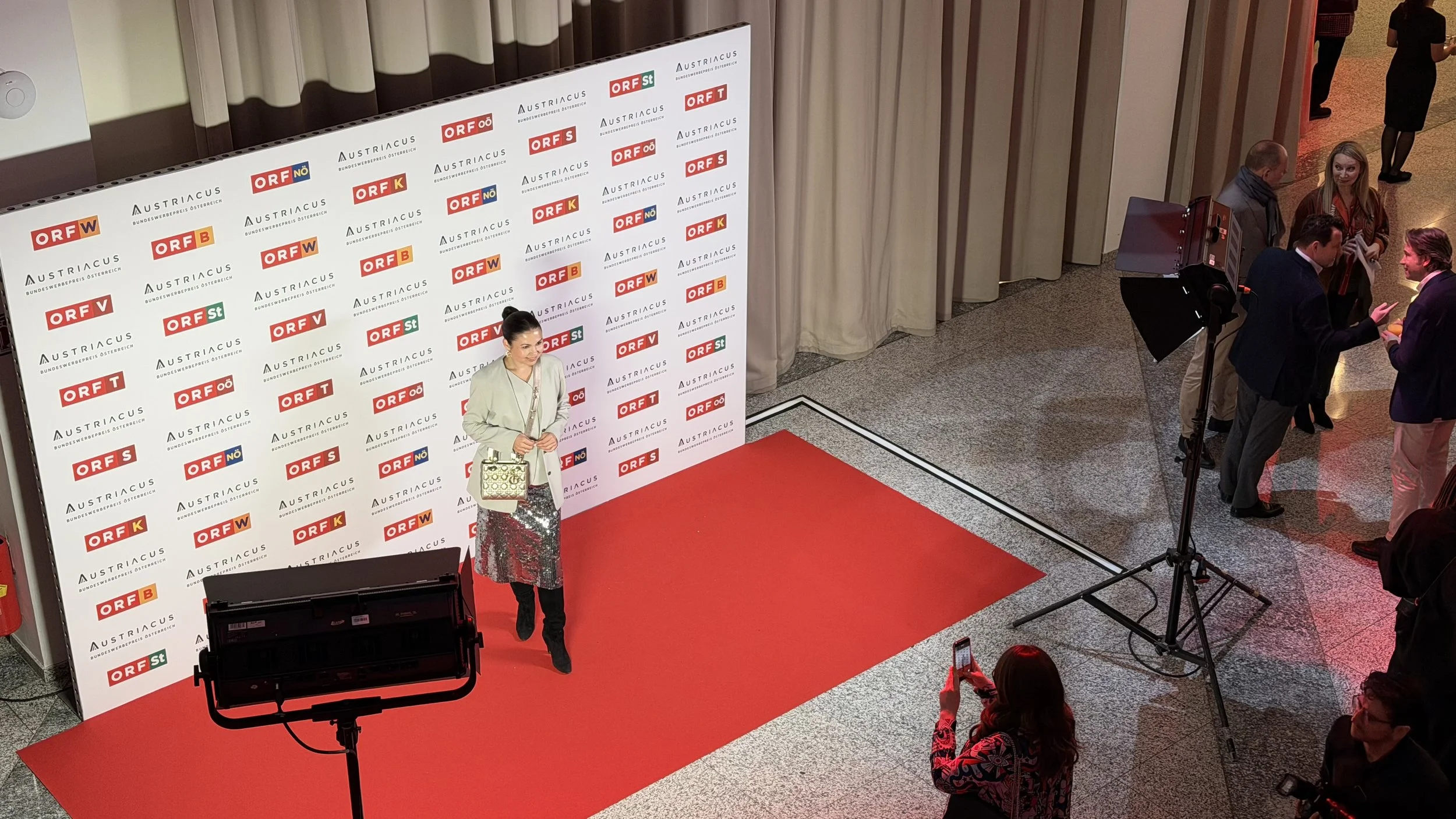 Eine Frau steht auf einem roten Teppich vor einem Hintergrund mit mehreren Logos und spricht in ein Mikrofon, während andere Personen im Hintergrund sich unterhalten oder Fotos machen.