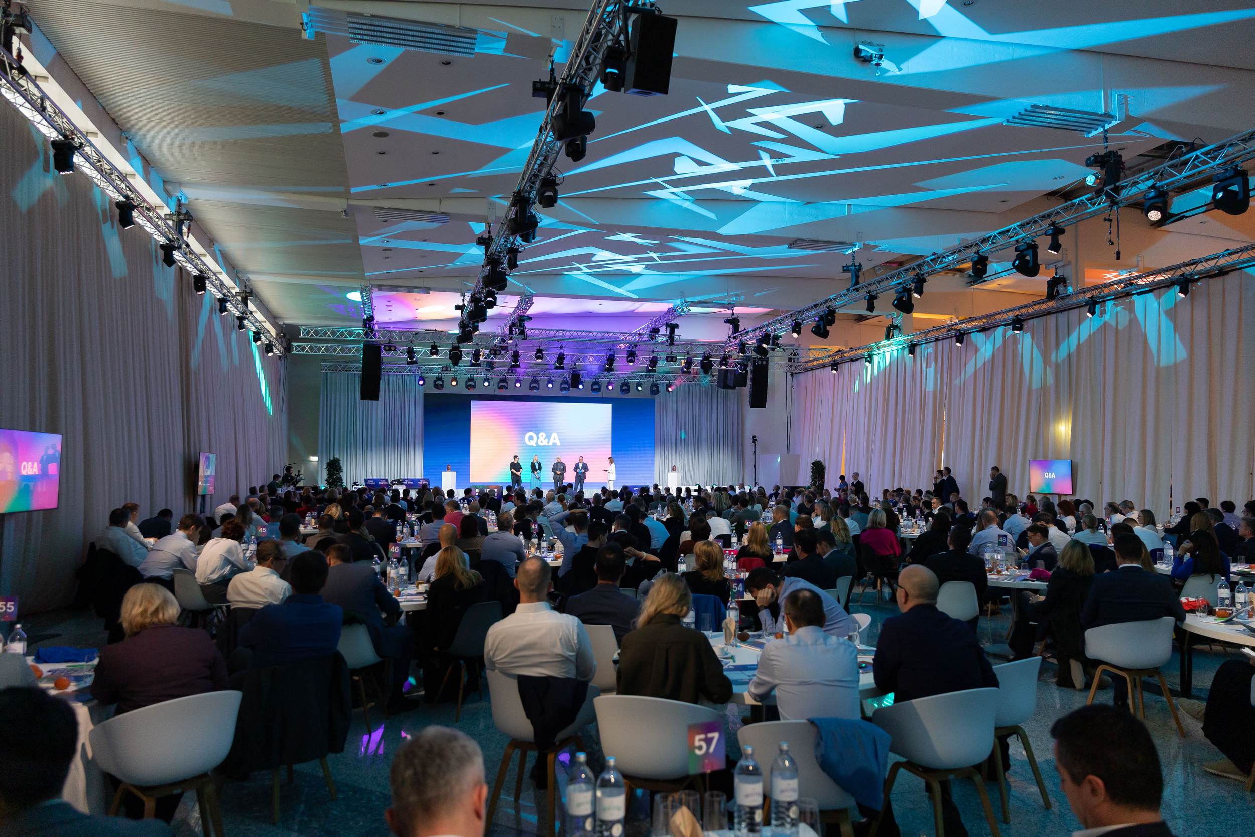 Eine große Konferenzhalle mit vielen Zuschauern, die einem Vortrag auf der Bühne bei einer Q&A-Session zuhören, mit mehreren Bildschirmen und bunter Beleuchtung.
