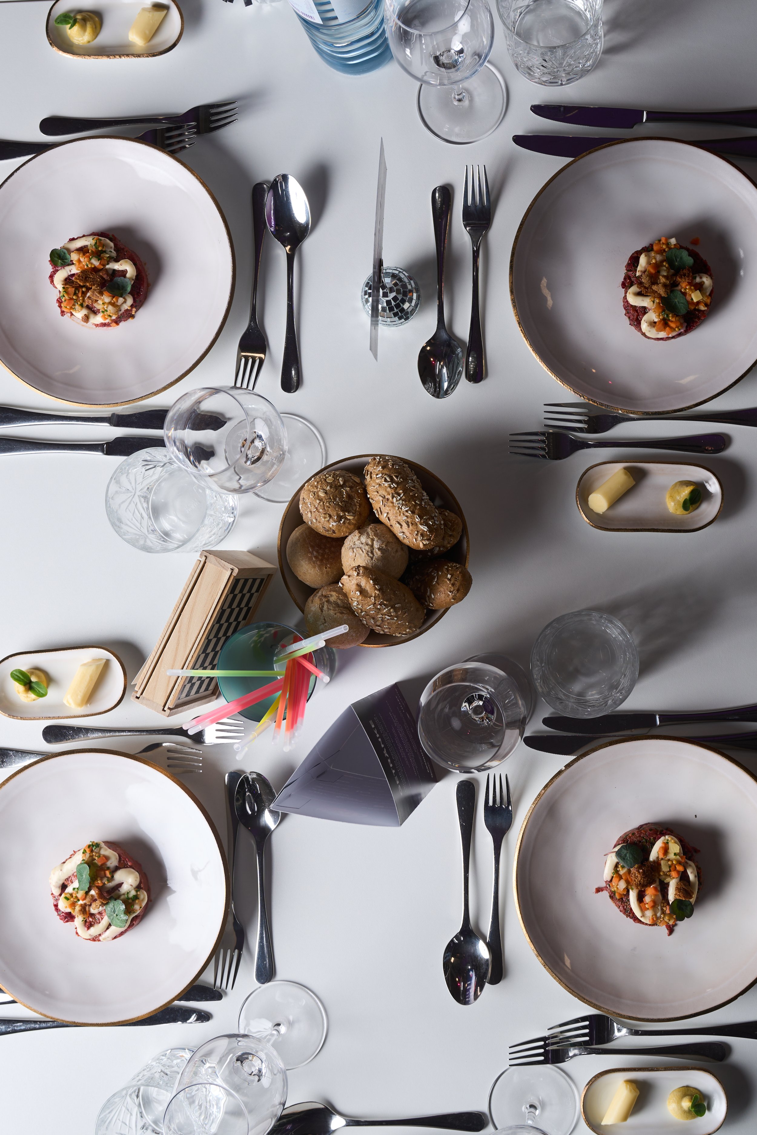 Tisch mit weißen Tellern, Gläsern, Besteck, Brot, und kleinen Desserts, für eine Dinner-Veranstaltung vorbereitet.