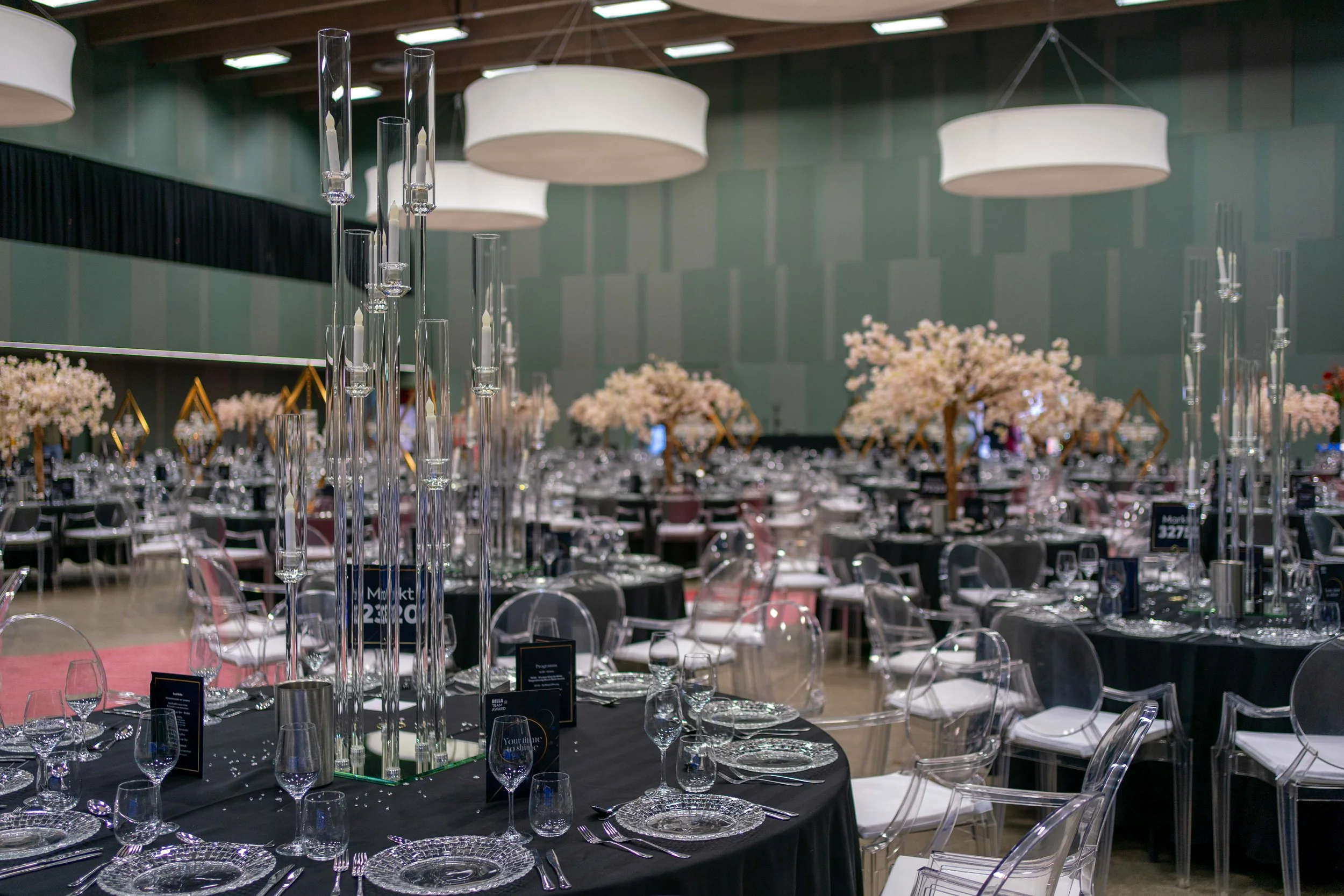 Eleganter Veranstaltungsraum mit mehreren runden Tischen, die mit schwarzen Tischdecken bedeckt sind, gehobene Kristallgläser, silberne Besteck, und zarte Blumenarrangements. Transparentes Stuhldesign und große weiße Hängelampen unterstreichen den fe