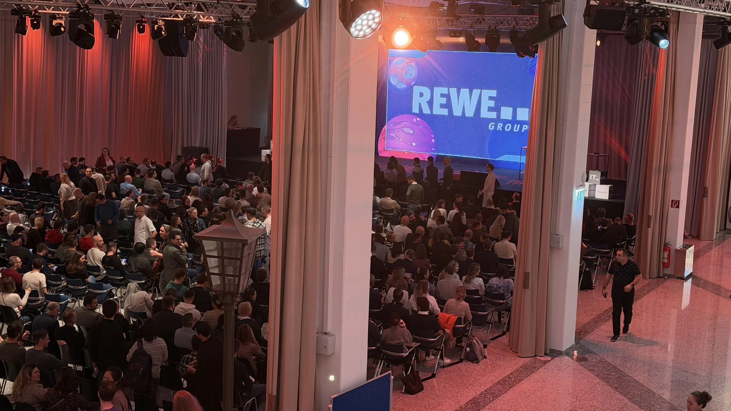 Innenaufnahme einer Veranstaltungshalle mit vielen Sitzplätzen, einem großen Bildschirm mit dem Logo der REWE Group, roten und weißen Vorhängen, sowie mehreren Personen, die sich auf die Veranstaltung vorbereiten.