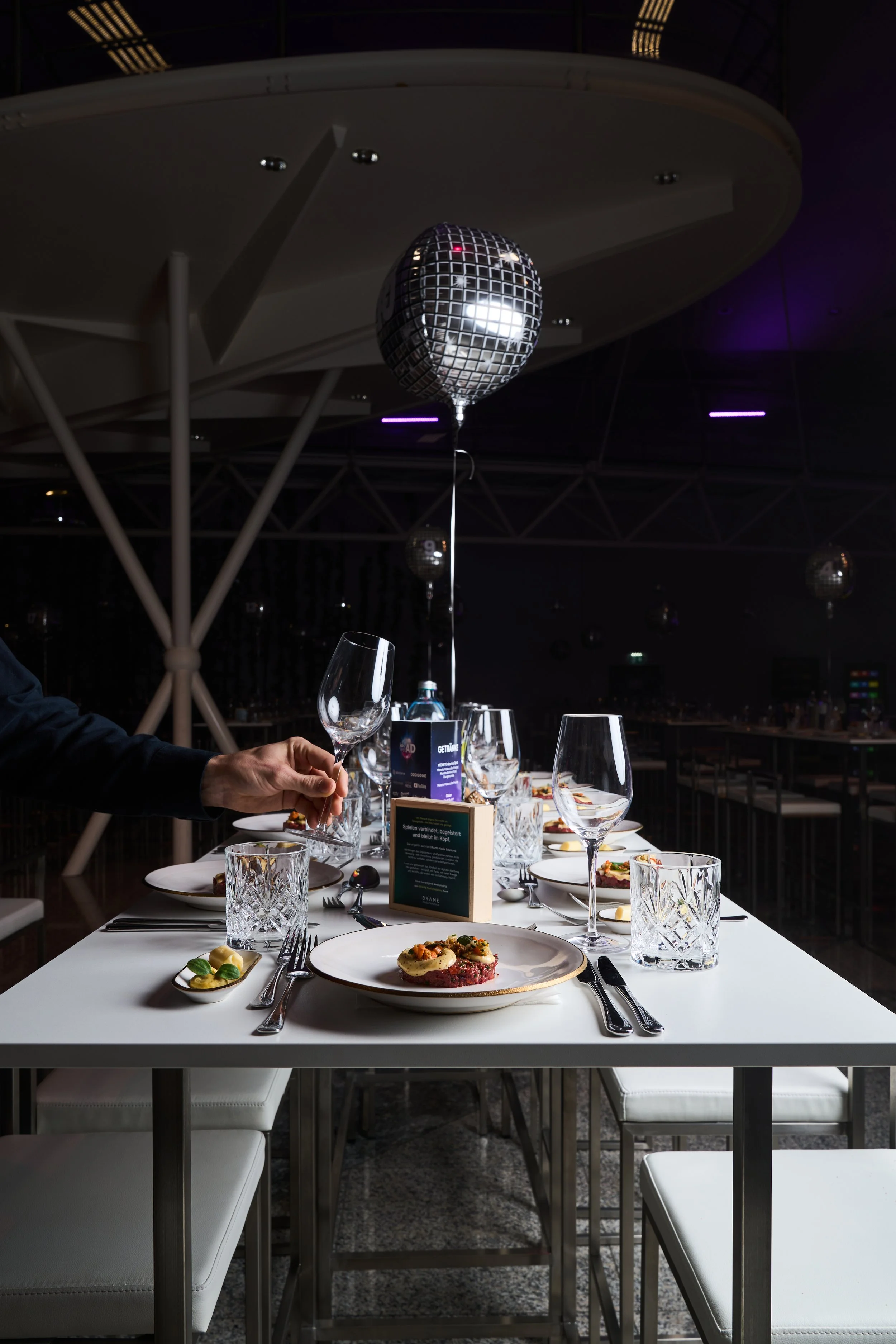 Ein festlich gedeckter Tisch mit leeren Weingläsern, Kristall-Gläsern, einem Teller mit Dessert, einem kleinen Snack, einem Menü und einer Hand, die ein Weinglas hält. Im Hintergrund Kugellampen und eine Disco-Kugel. 