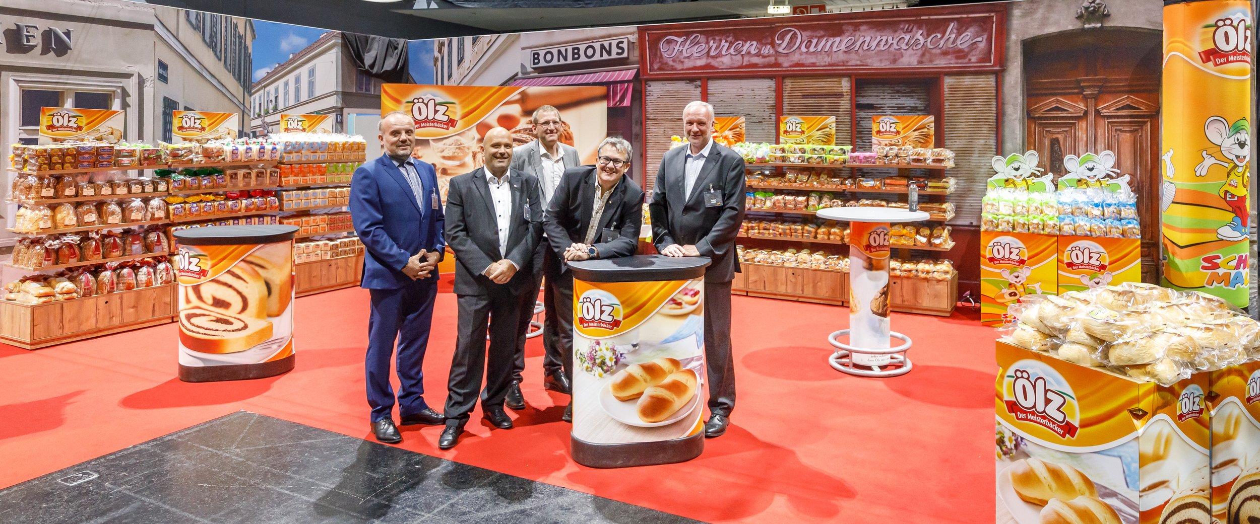 Fünf Männer in formeller Kleidung stehen in einer Ausstellungsfläche für Backwaren, umgeben von Regalen mit Brötchen, Süßigkeiten und Plakaten, im Hintergrund eine Kulisse mit Stadtansicht und Geschäftsschildern.