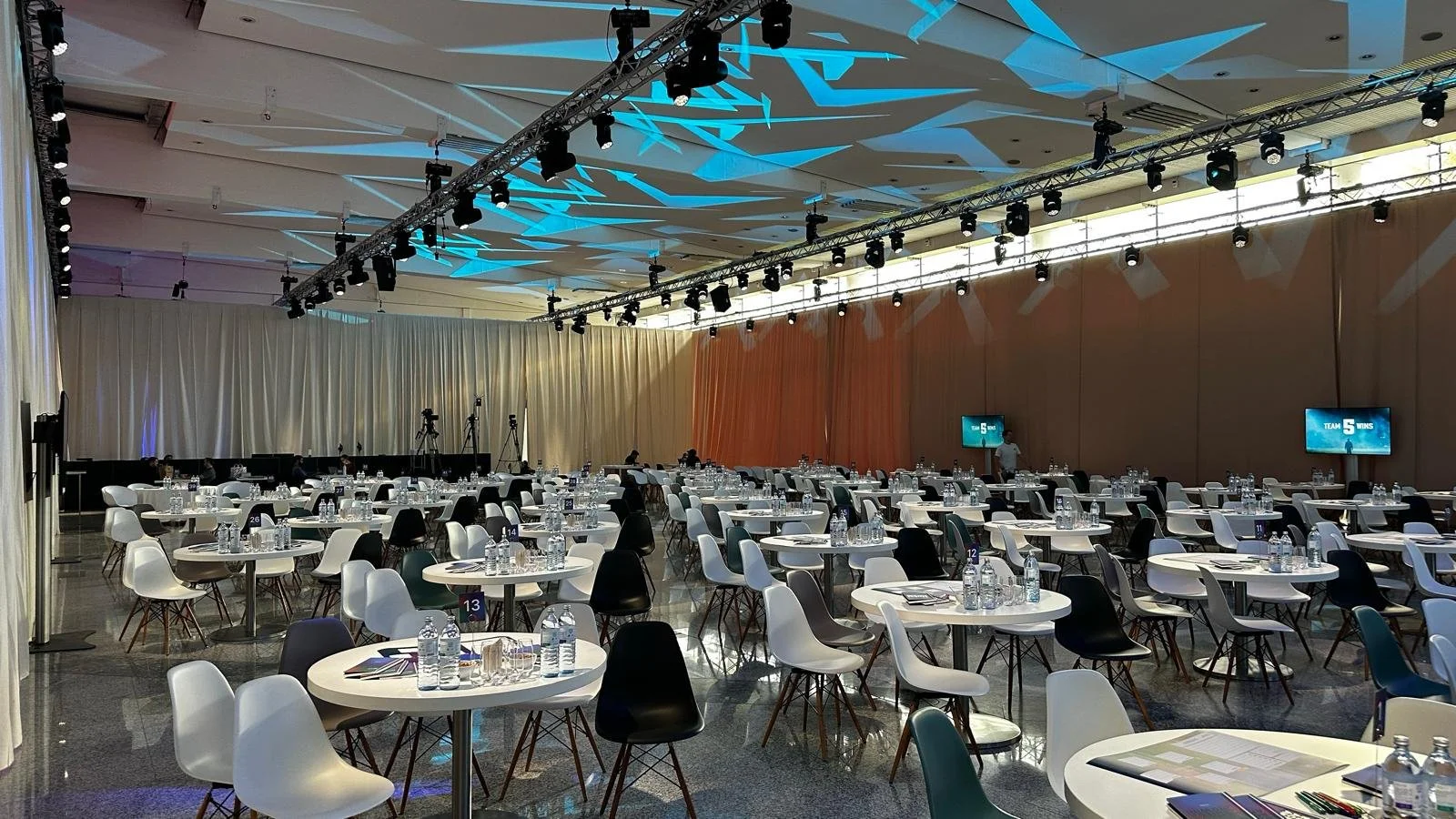 Leerer Konferenzsaal mit runden Tischen, Stühlen, Wasserflaschen, Schreibmaterialien und Präsentationsbildschirmen, vorbereitet für ein Event.